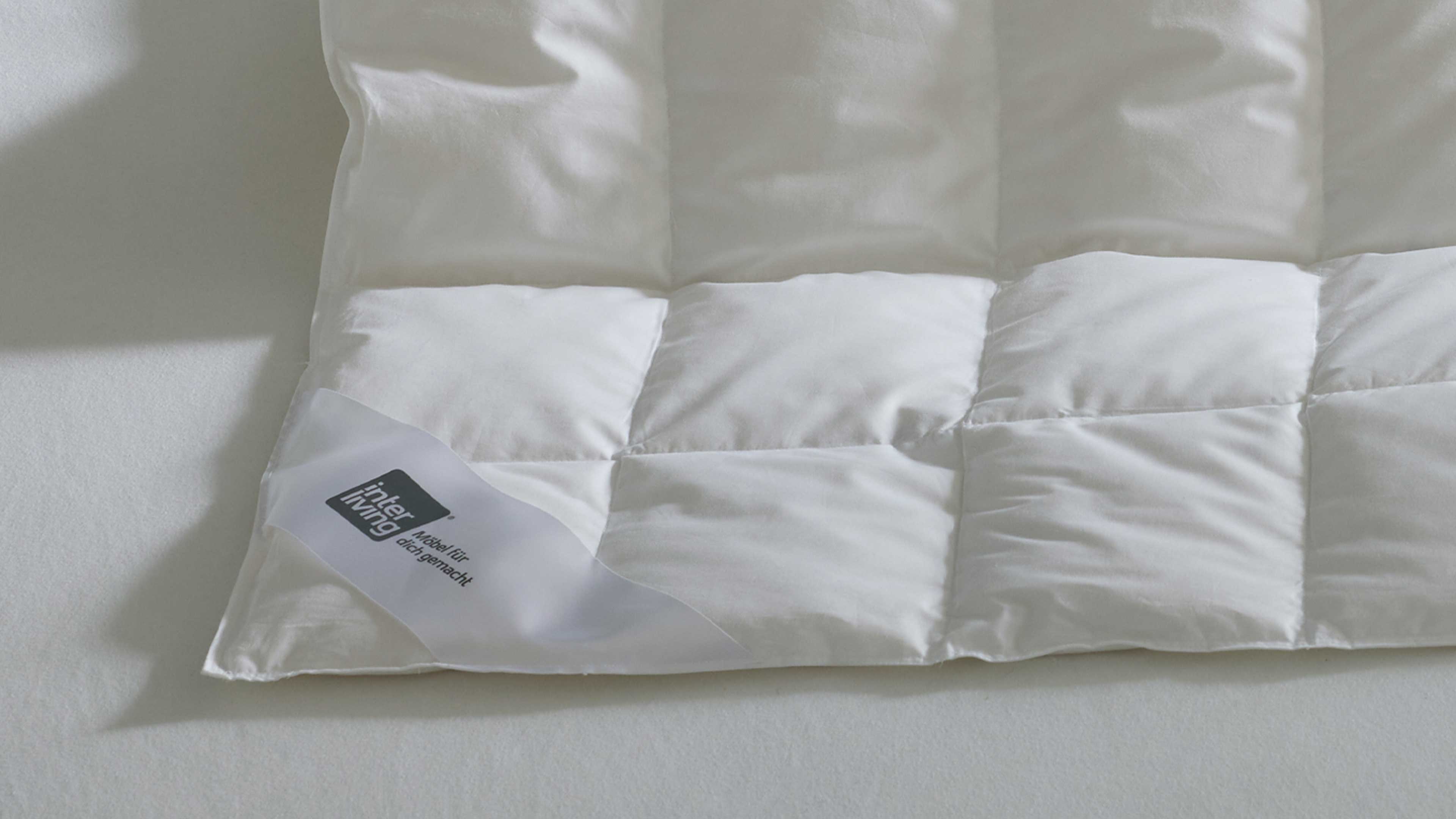 Gesteppte weiße Bettdecke auf hellem Untergrund, sichtbare Ecke mit weißem Etikett und schwarzem Schriftzug 'interliving'.