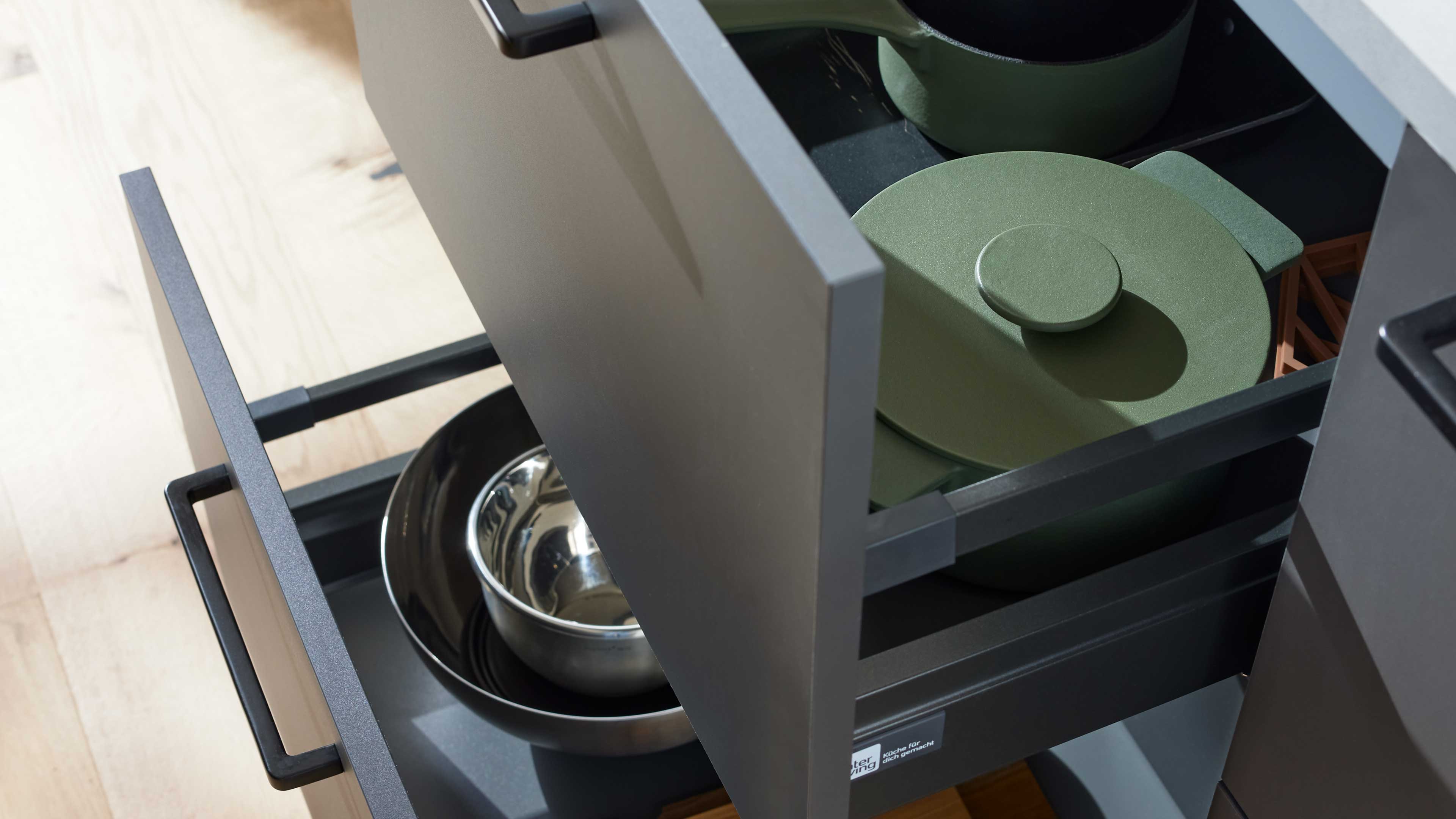 Offene graue Küchenschubladen mit schwarzem Griff, darin grüner Kochtopf mit Deckel sowie ineinander gestapelte Edelstahl- und Pfannen.