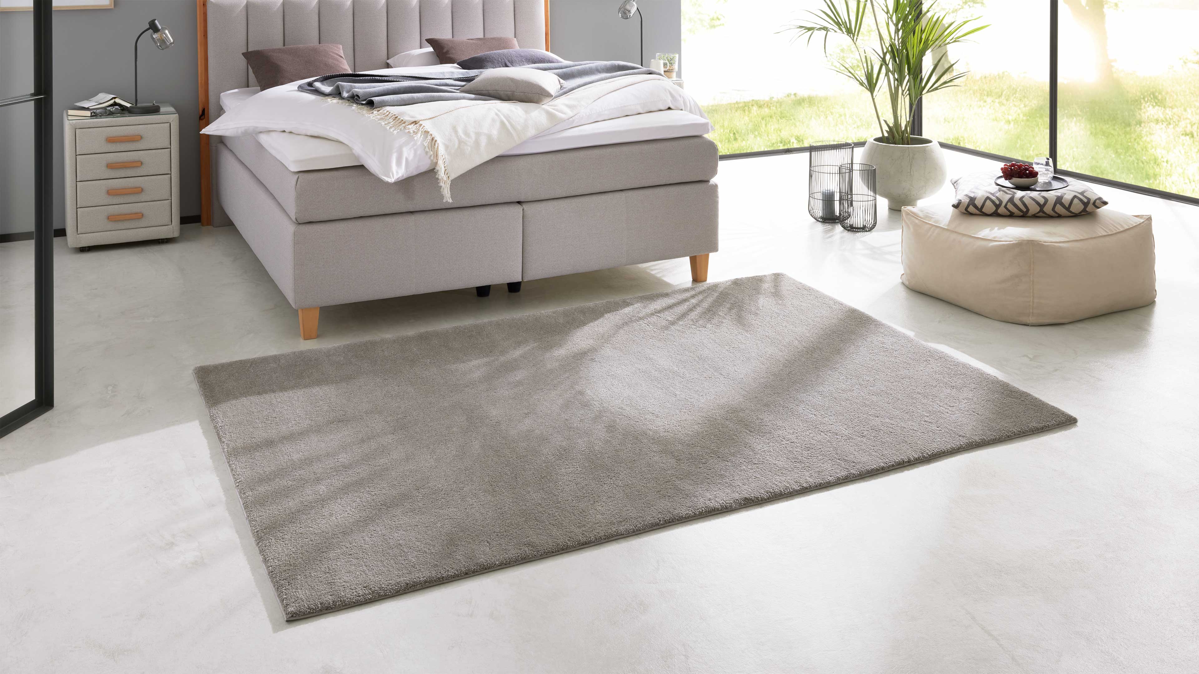 Grau bezogenes Doppelbett mit gepolstertem Kopfteil, weiße Bettwäsche und Decke, daneben Nachttisch, Teppich, Pouf und Pflanze.