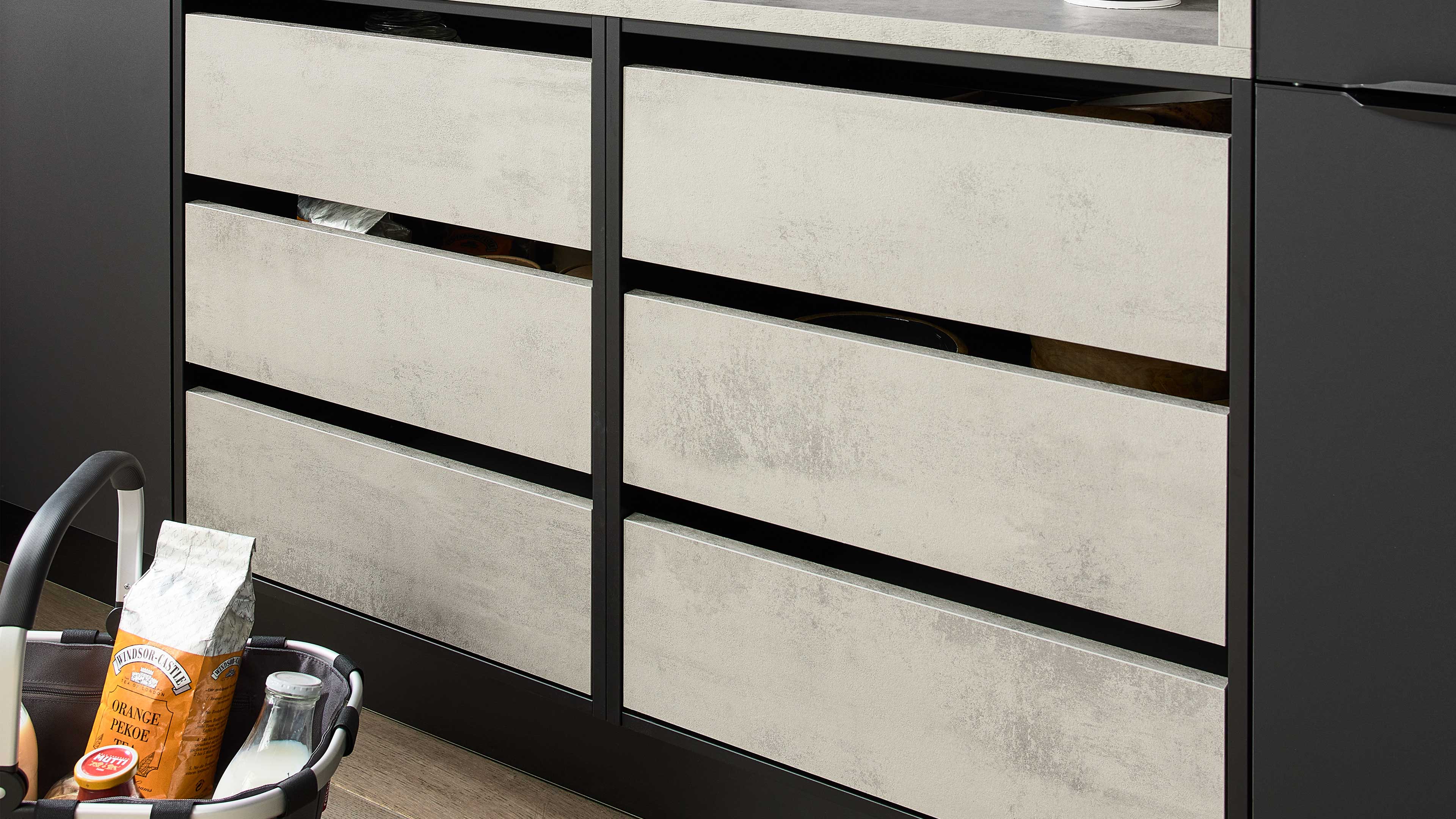 Zweispaltiger Schubkastenschrank mit sechs geöffneten hellgrauen Betonoptik-Fronten im schwarzen Rahmen und Einkaufskorb links unten.