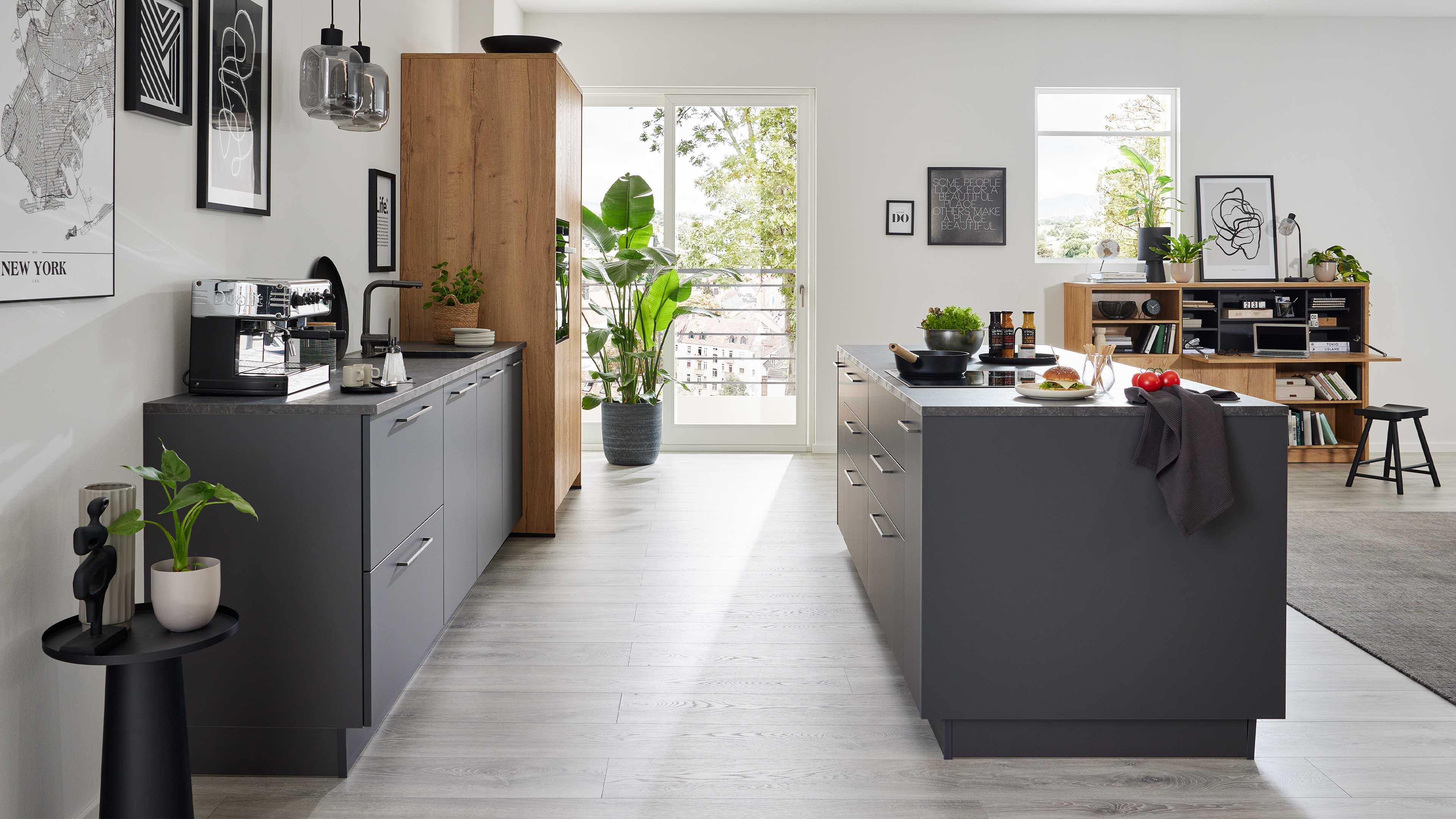 Offene Küche mit zwei dunkelgrauen Arbeitszeilen, Insel, Hochschrank aus Holz, Kaffeemaschine und Topfpflanze vor Terrassentür.