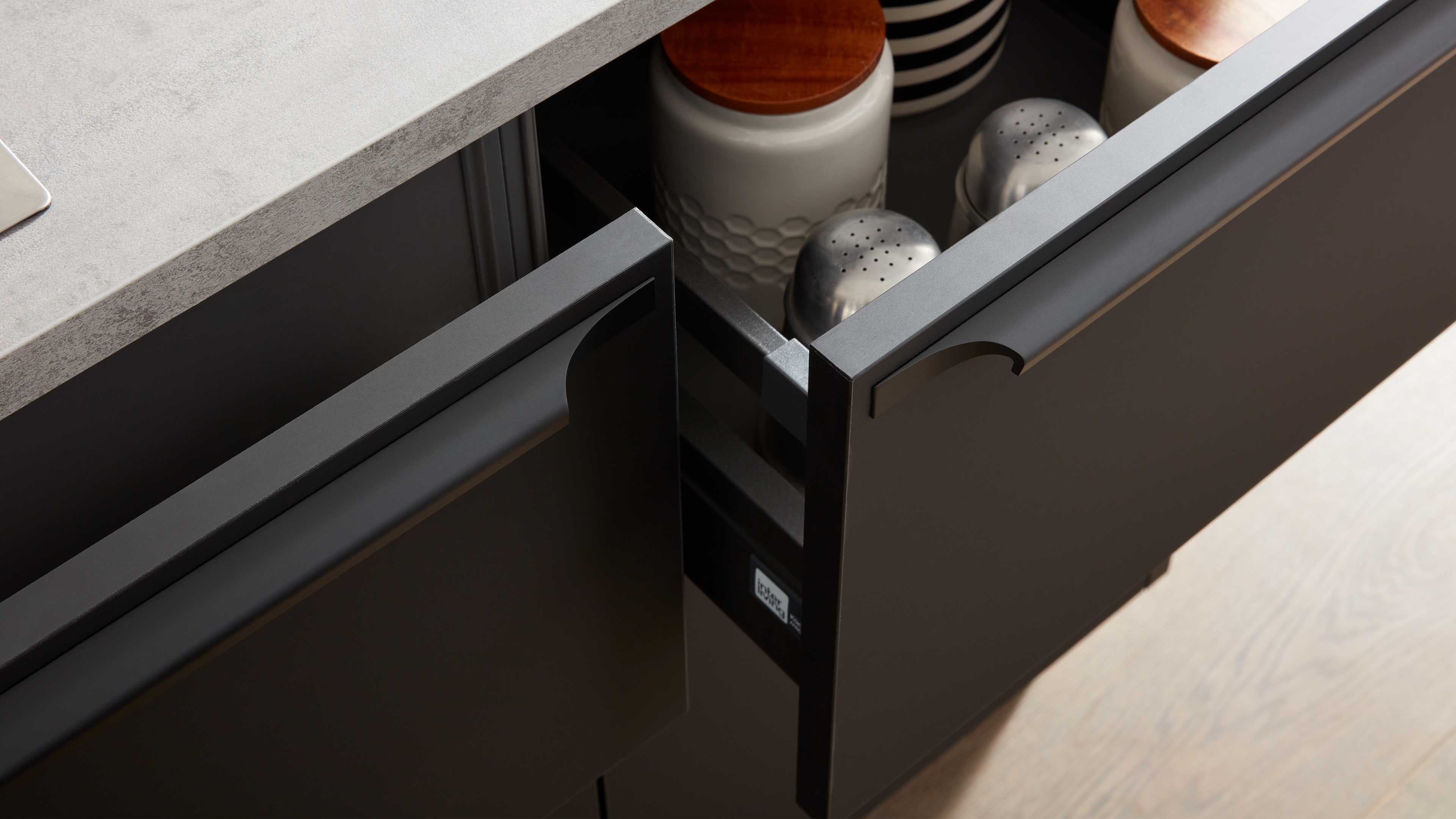 Zwei halb geöffnete dunkelgraue Küchenschubladen unter hellem Arbeitsplattenrand; innen Keramikdosen mit Holzdeckeln und Metallstreuern.