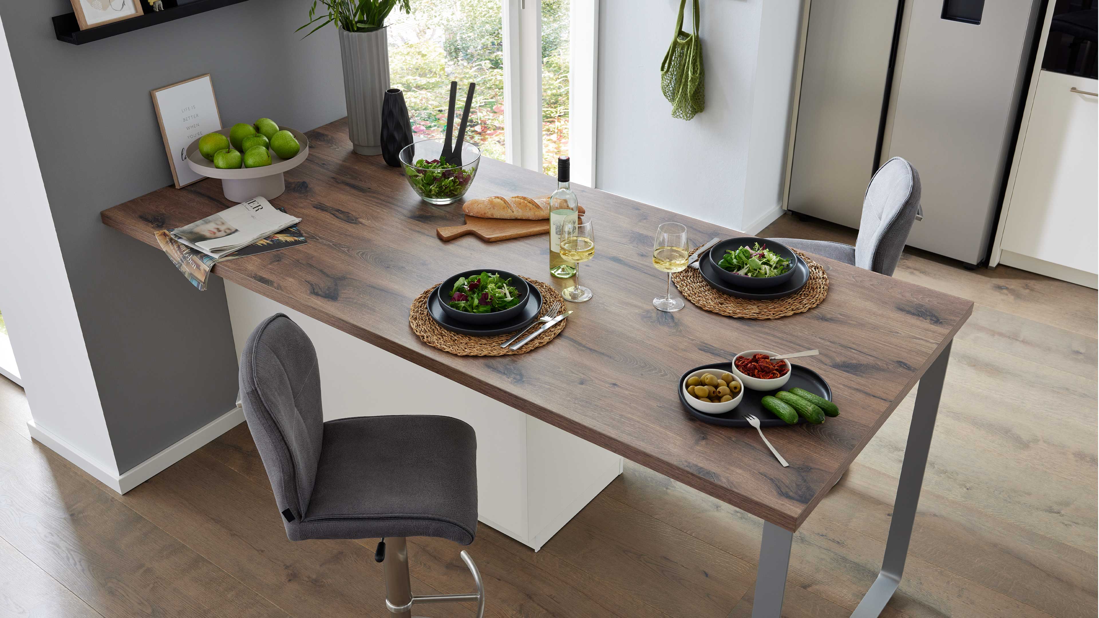 Rechteckige Holzarbeitsplatte auf Metallbeinen, zwei graue Stühle, gedeckte Salatteller, Weinflasche, Brot, Schalen mit Oliven und Gurken.