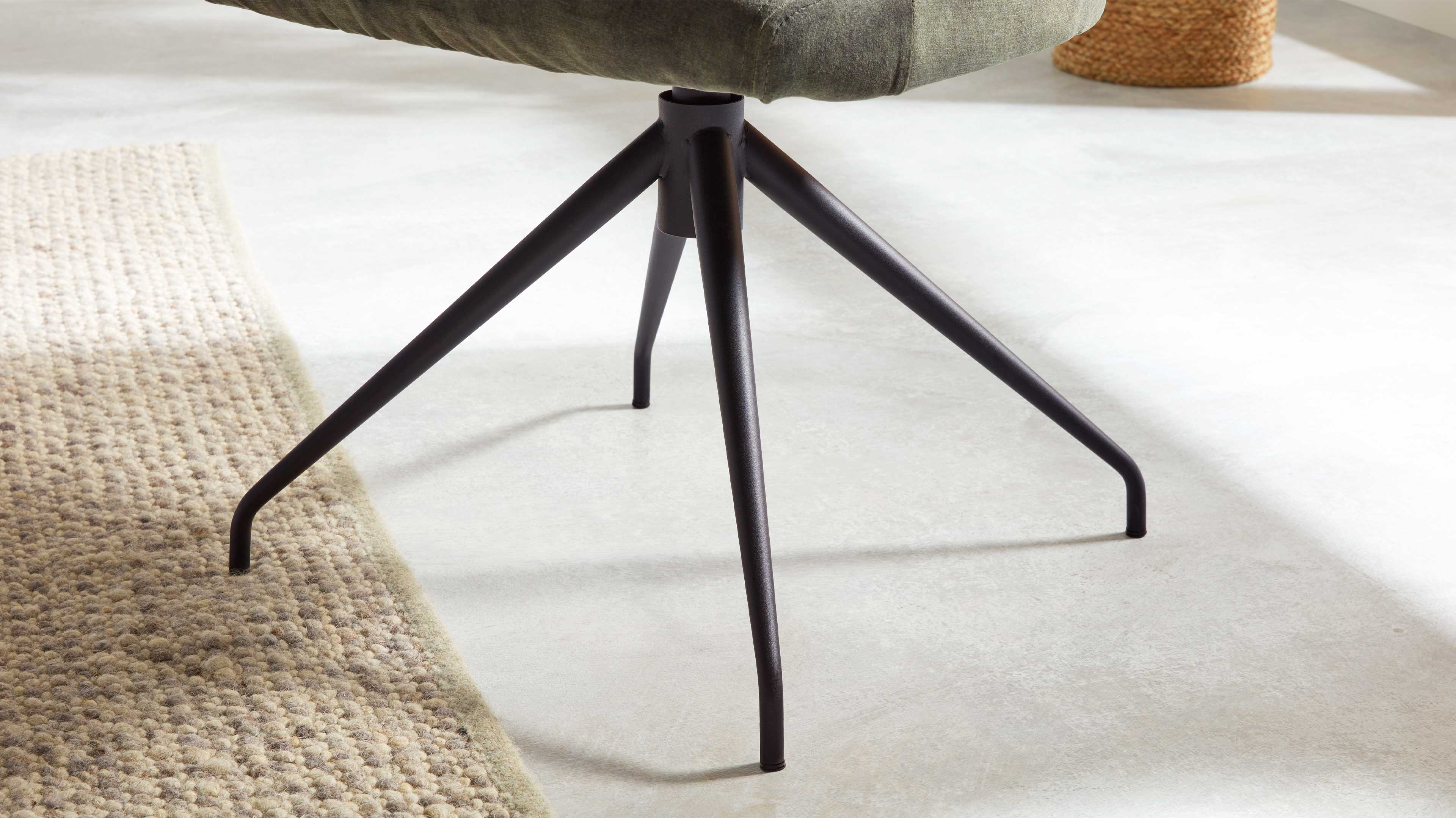 Schwarze, vierarmige Metallfußkreuz eines Drehstuhls mit grünem Polsterrand, auf hellem Fußboden neben einem strukturierten Teppich.