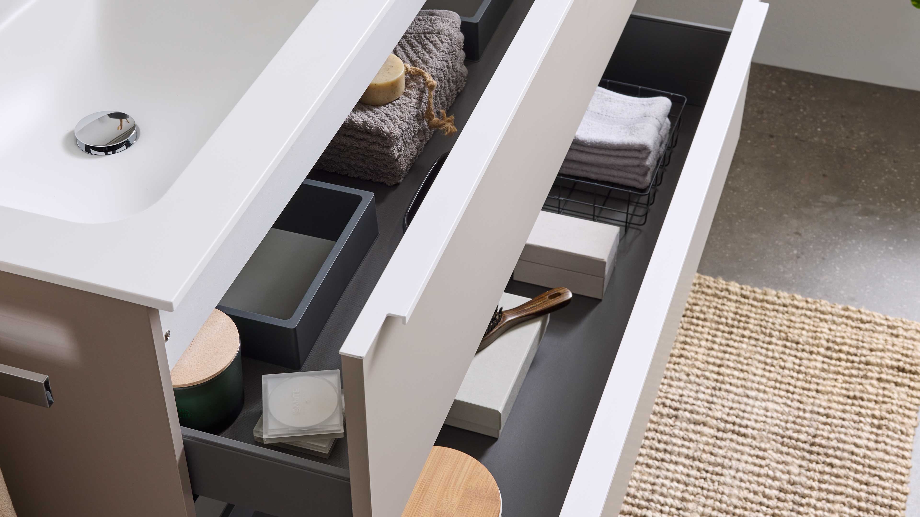 Geöffnete weiße Waschtischschublade mit grauem Innenraum, gefalteten Handtüchern, Seife, kleinen Boxen und einer Holzbürste.