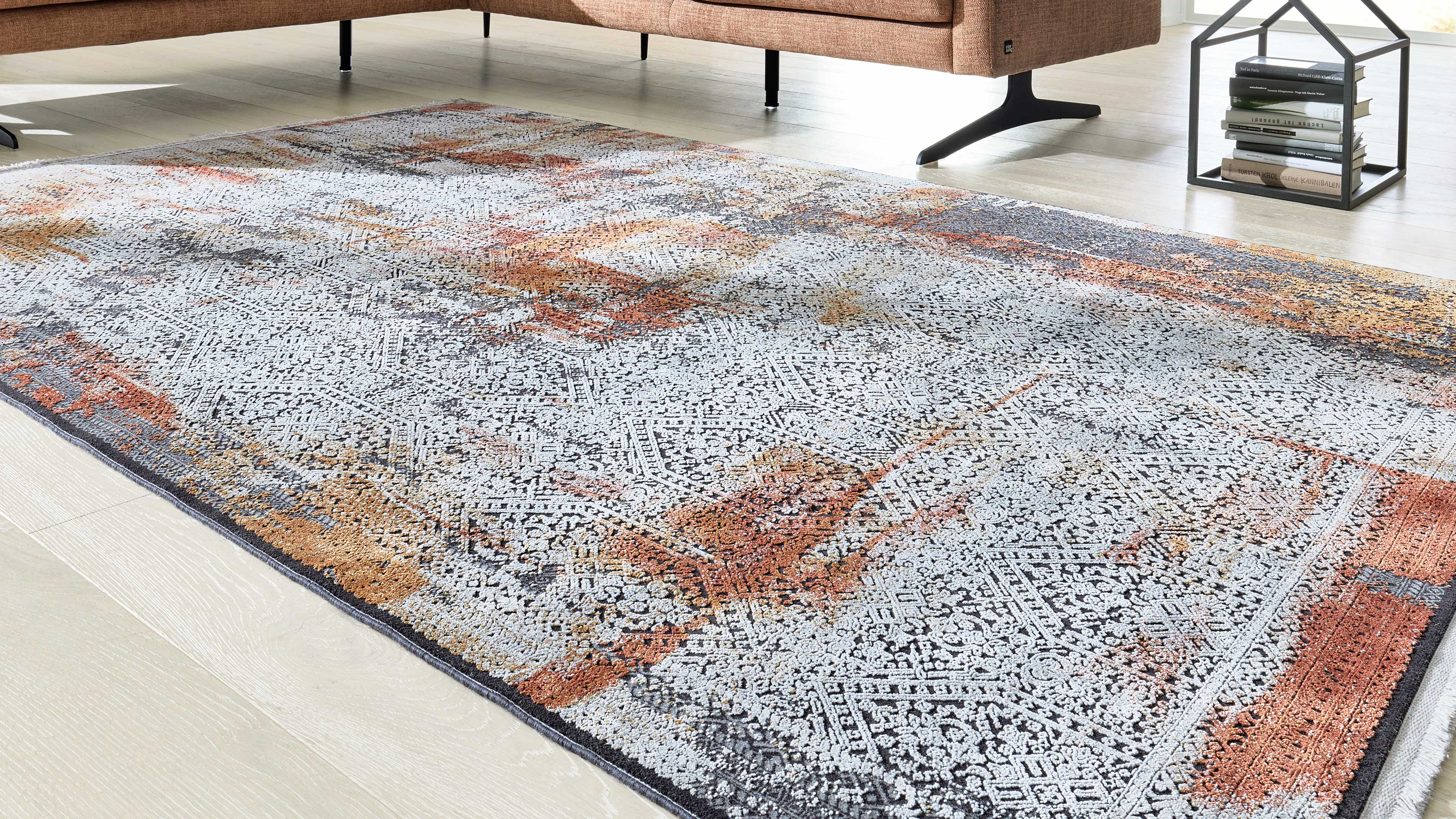 Großflächiger hellgrau-weiß gemusterter Teppich mit rost- und goldfarbenen Akzenten, im Hintergrund Sofa und offenes Metallregal mit Büchern.