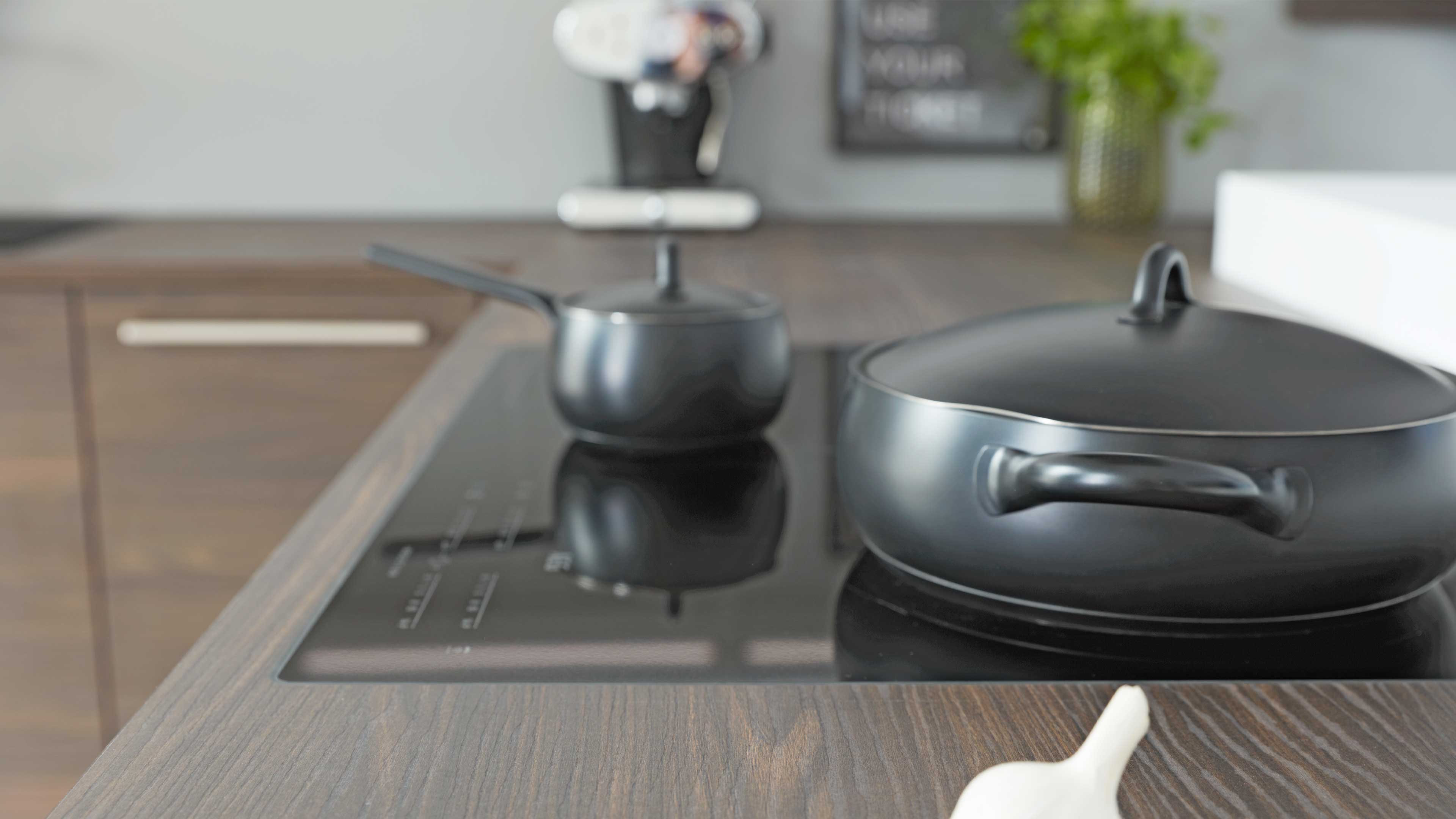 Schwarzes Glaskochfeld in dunkler Holzarbeitsplatte mit zwei mattschwarzen Töpfen, einem flachen Bräter mit Deckel und einem kleinen Stieltopf.