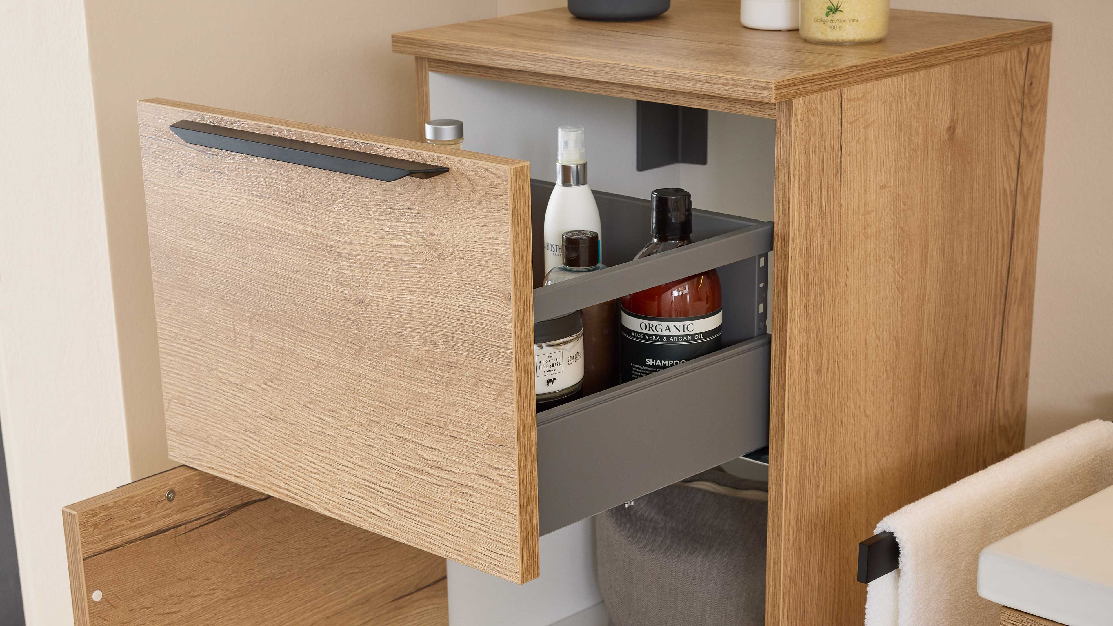 Offene Holzschublade mit grauem Inneneinsatz, mehreren Pflegeflaschen und -tiegeln, rechts ein aufgehängtes Handtuch.