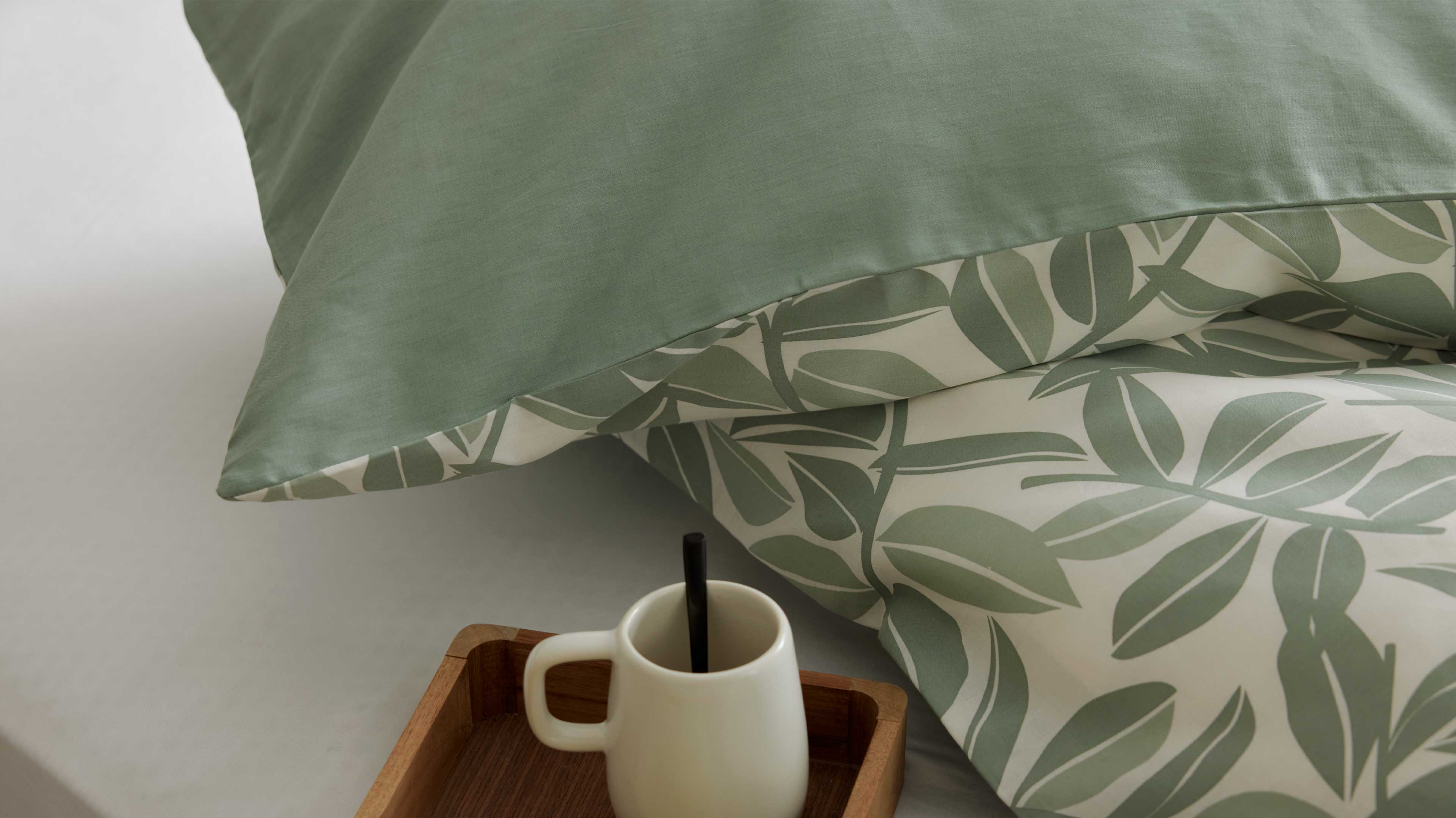 Zwei Kissen in Grün und mit grünem Blattmuster liegen übereinander, dazu ein Holztablett mit weißer Tasse und schwarzem Strohhalm.