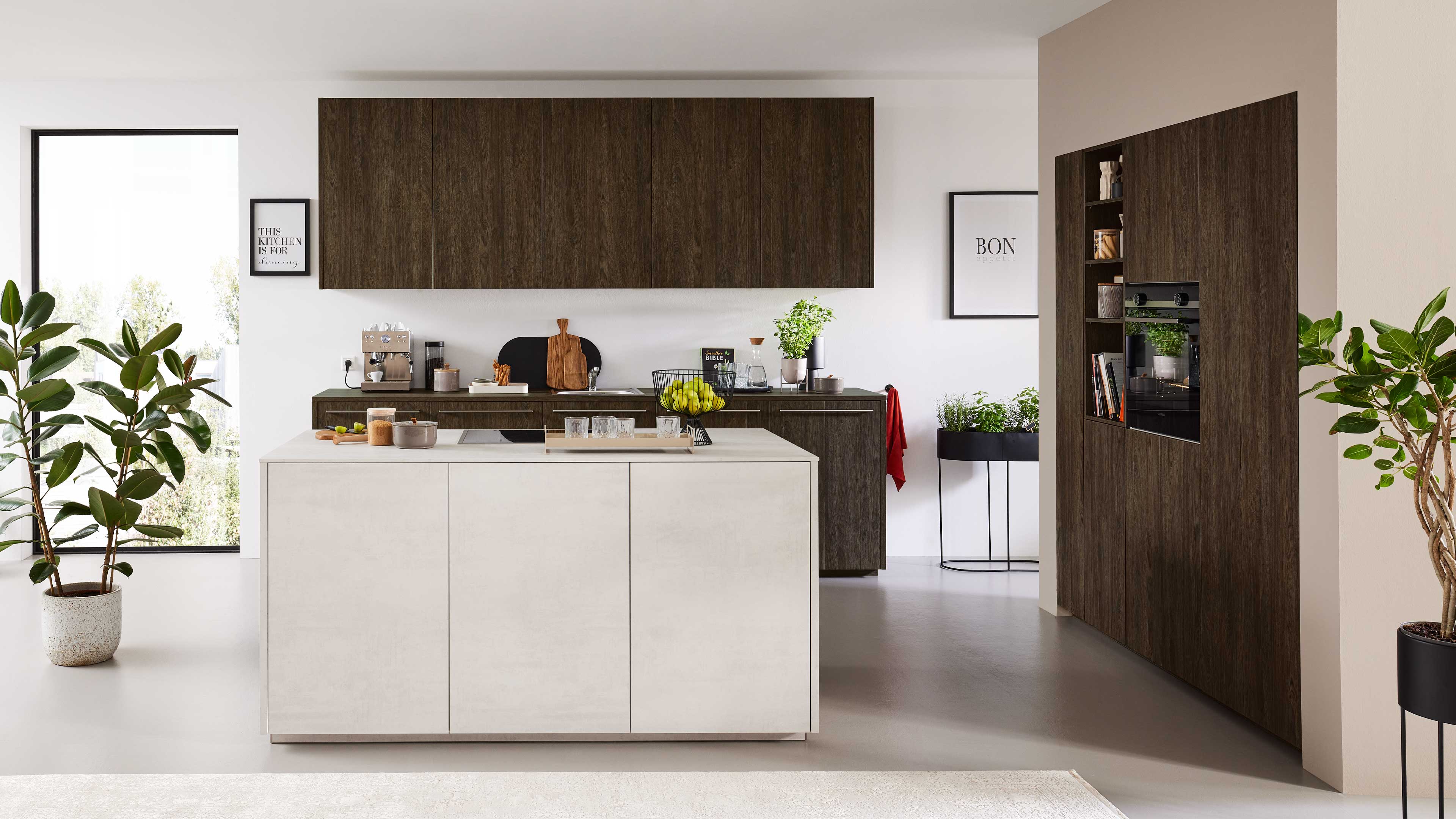 Helle Kücheninsel im Vordergrund mit Tablett und Schale mit Bananen, dahinter dunkelbraune Holzfronten, Einbaugeräte und Zimmerpflanzen.