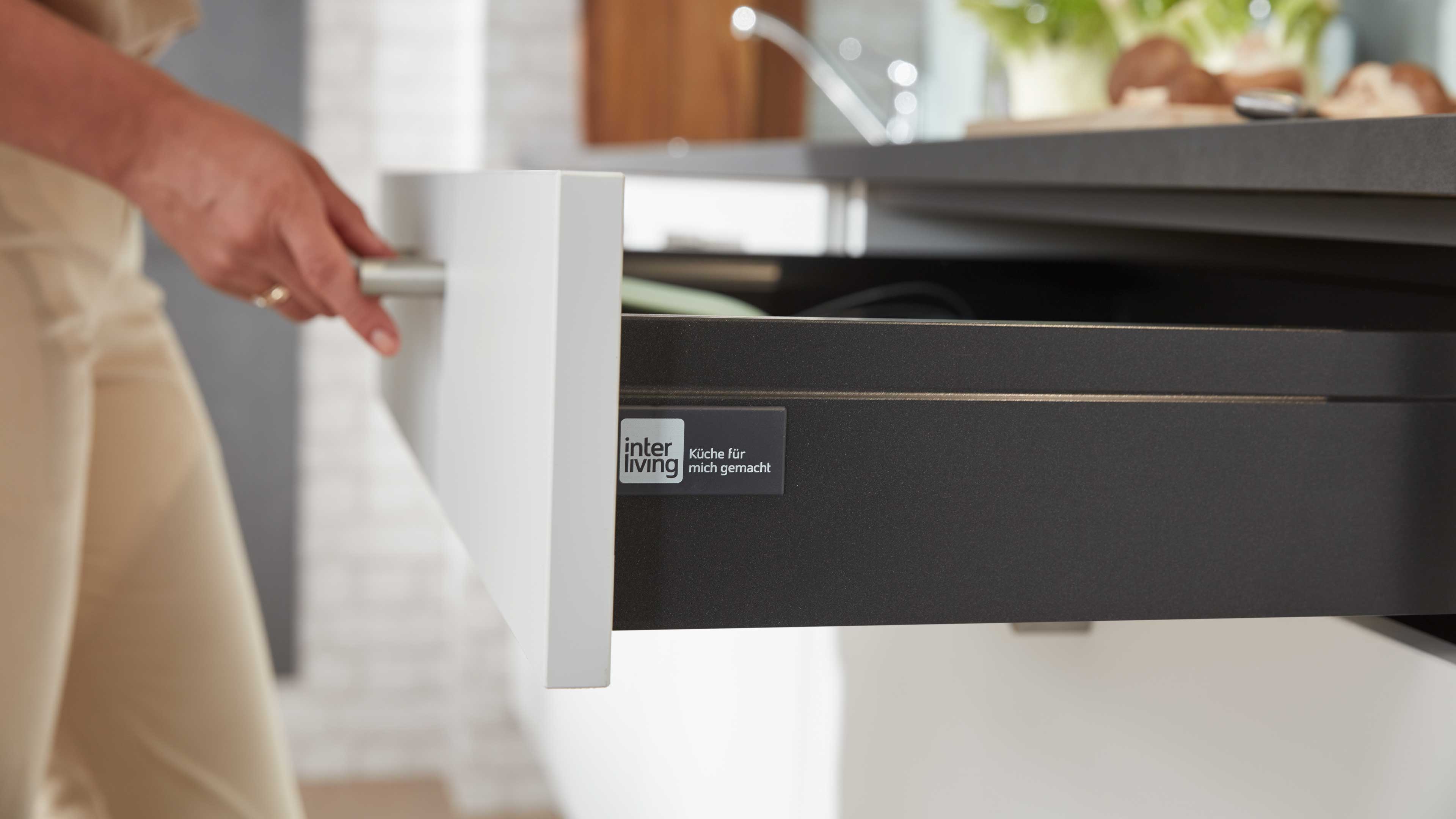Geöffnete weiße Küchenschublade mit rundem Metallgriff, dunkle Innenfront des Schubkastens mit sichtbarem "interliving"-Aufkleber.