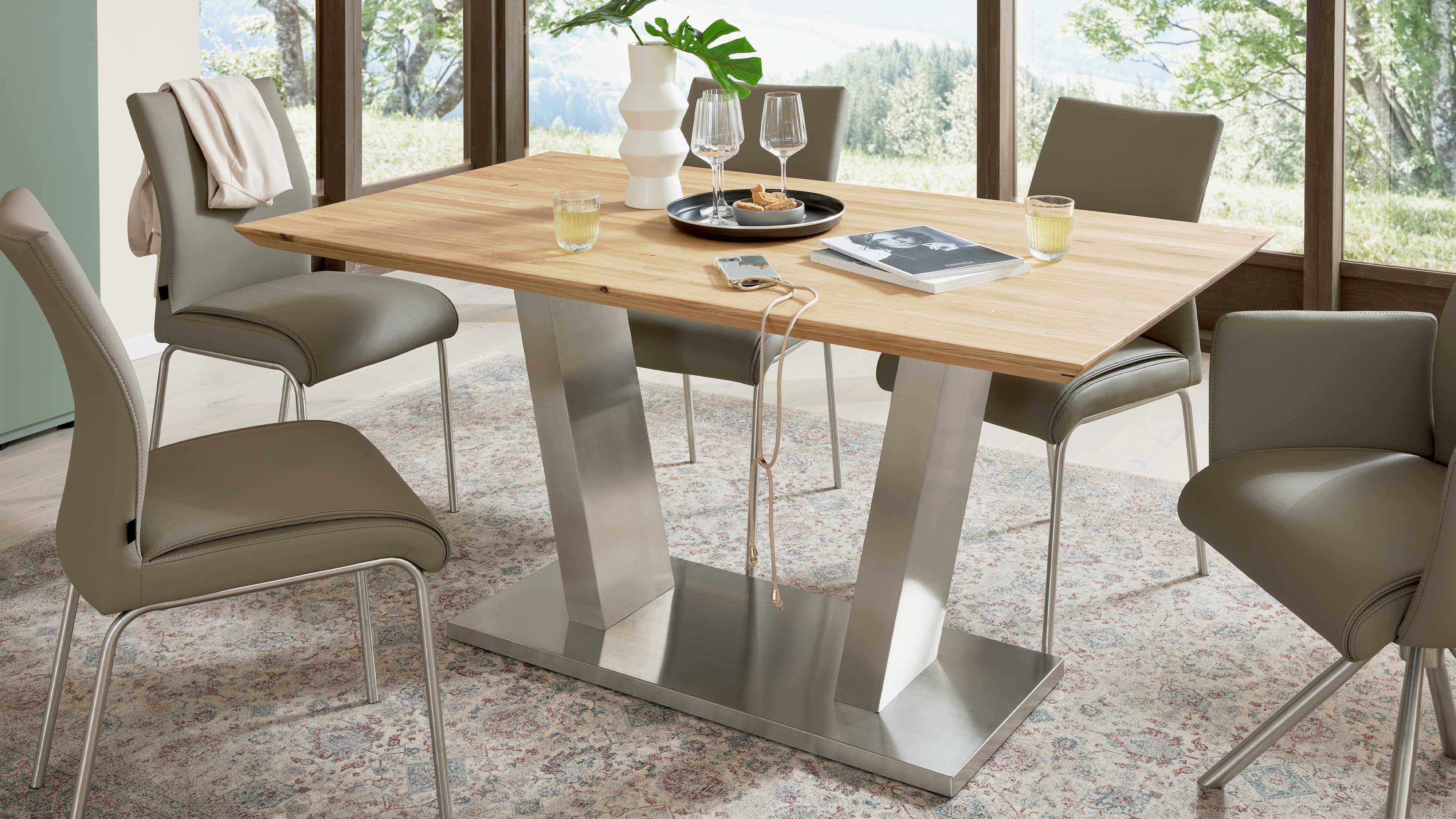 Holztisch mit rechteckiger Platte auf zwei V‑Metallstützen, umgeben von grauen Stühlen und mit Vase, Gläsern, Tablett und Buch.