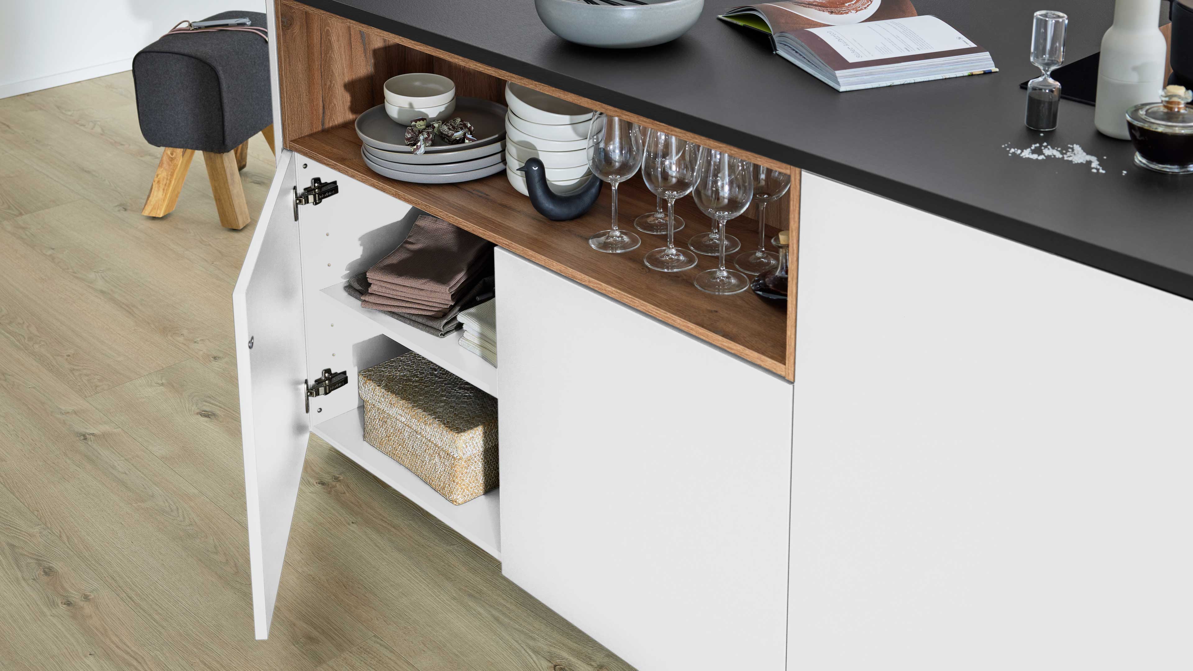 Weißer Unterschrank mit schwarzer Arbeitsplatte und offenem Fach mit Tellern, Weingläsern, gefalteten Tüchern sowie gewebter Box.