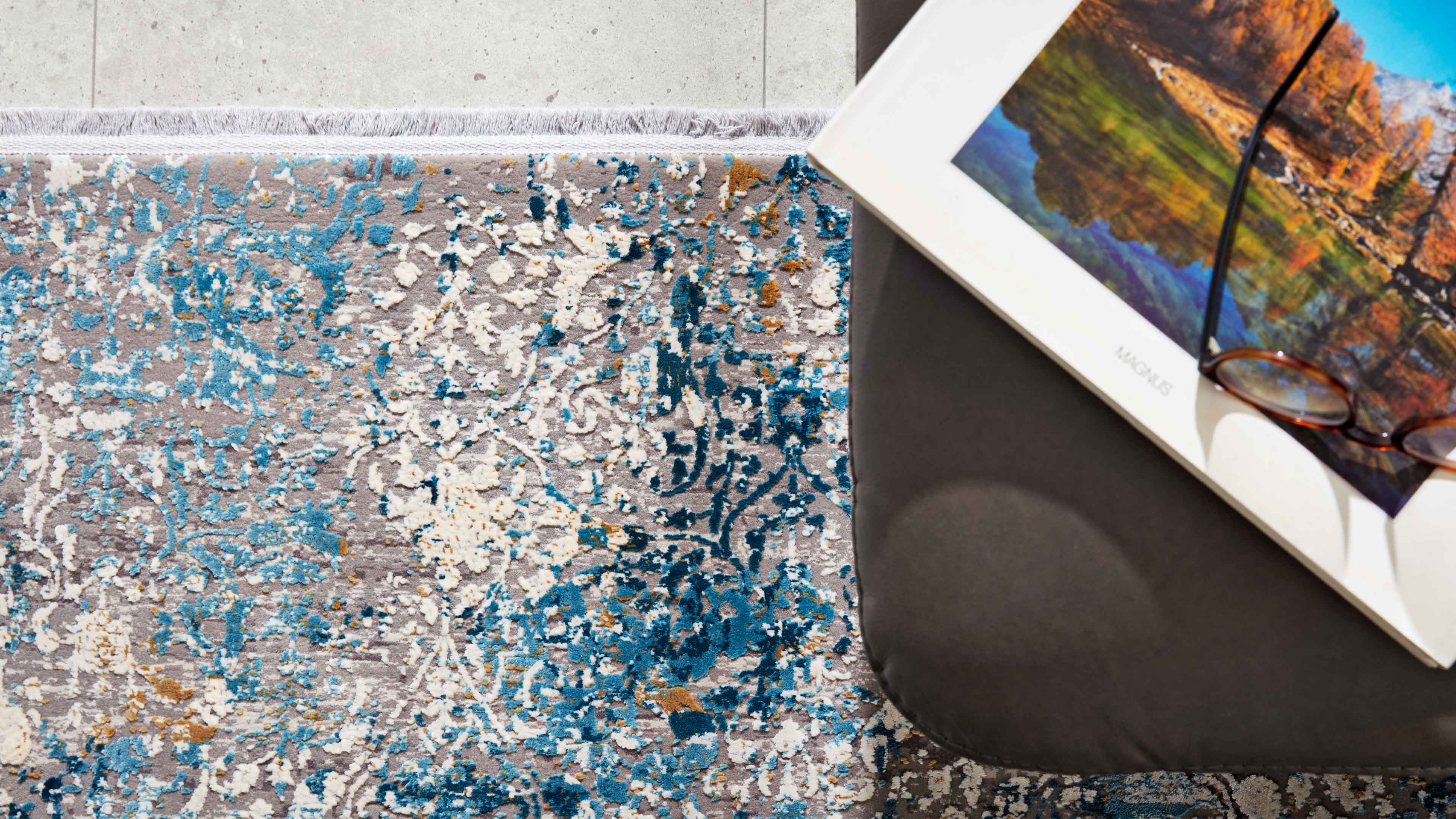 Grauer Teppich mit blau-weißen und goldenen Ornamenten und sichtbaren Fransen, rechts ein dunkles Polster mit gerahmtem Bild und Brille.