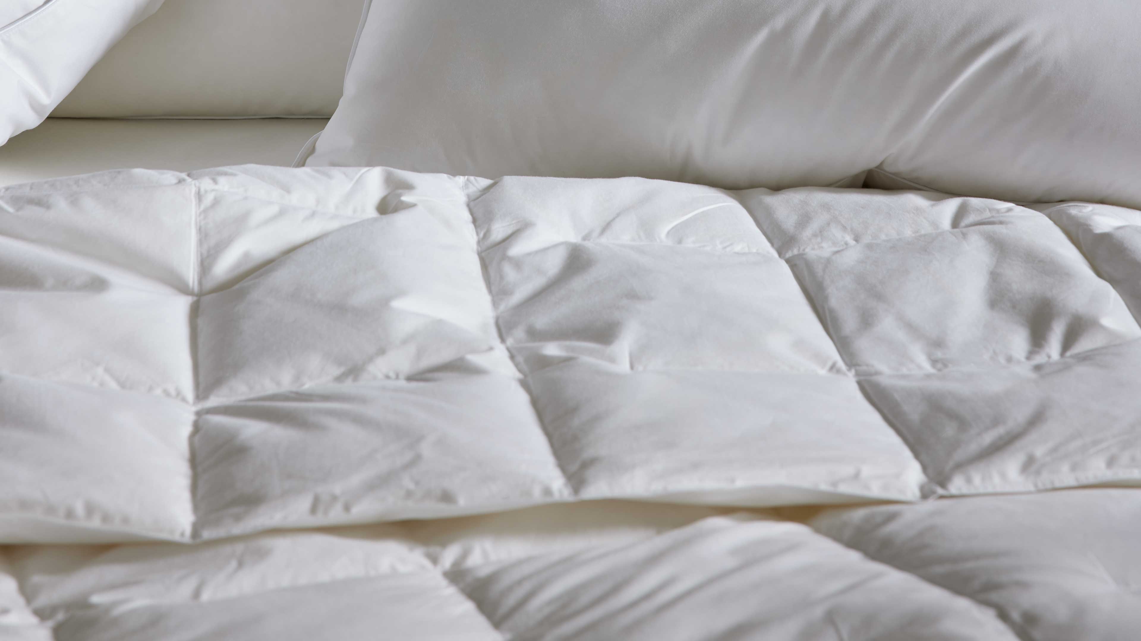 Weiß gestepptes Bettdeckenfeld mit quadratischen Kammern liegt auf dem Bett, im Hintergrund weiße Kopfkissen.