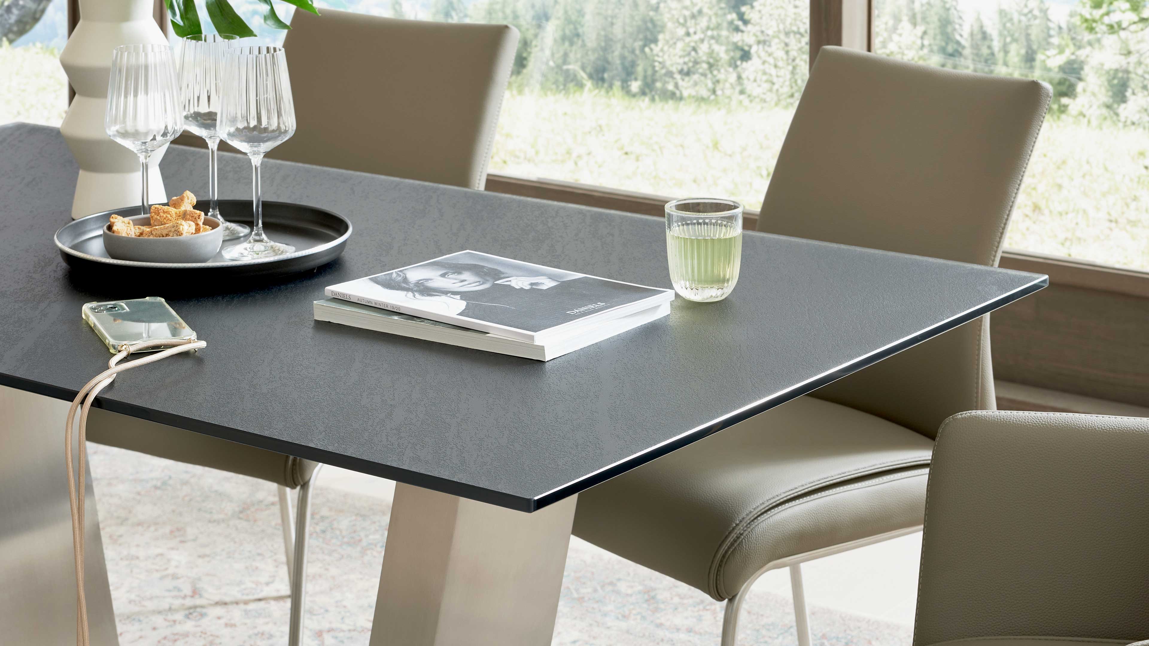 Dunkler rechteckiger Esstisch mit strukturierter Oberfläche, beigefarbenen Polsterstühlen, Magazin, Trinkglas und Tablett mit Weingläsern.