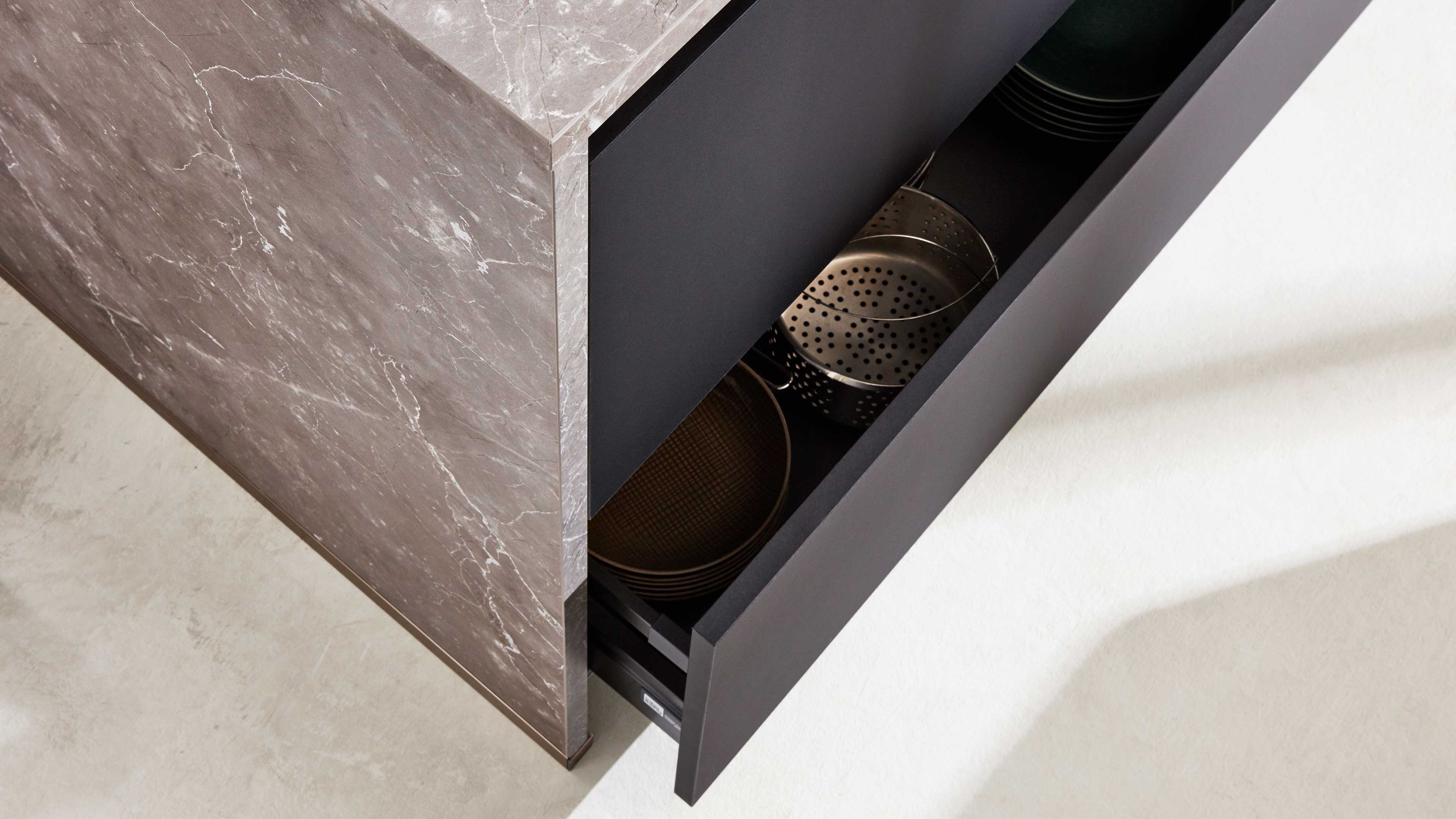 Ausgezogene schwarze Schublade an Insel mit graubrauner Marmorfront, darin gelochener Metallsieb und gestapelte runde Küchengegenstände.