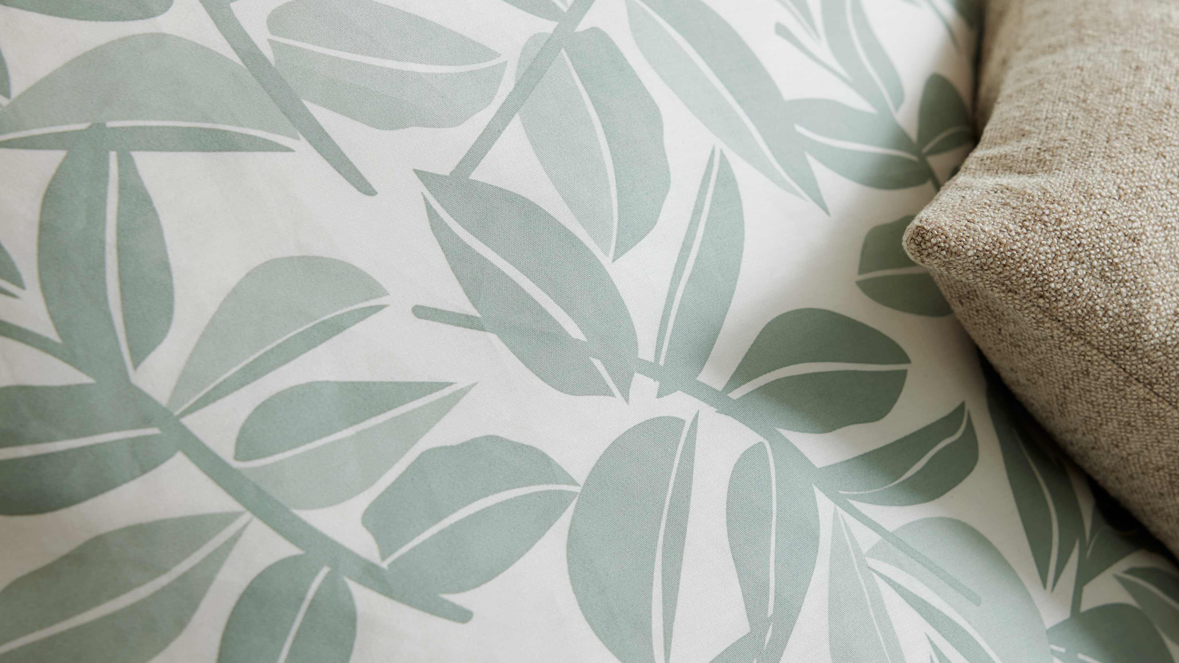 Großformatiges Stoffmuster mit hellgrünen, stilisierten Blättern auf weißem Hintergrund, rechts ein beiges Strukturkissen.