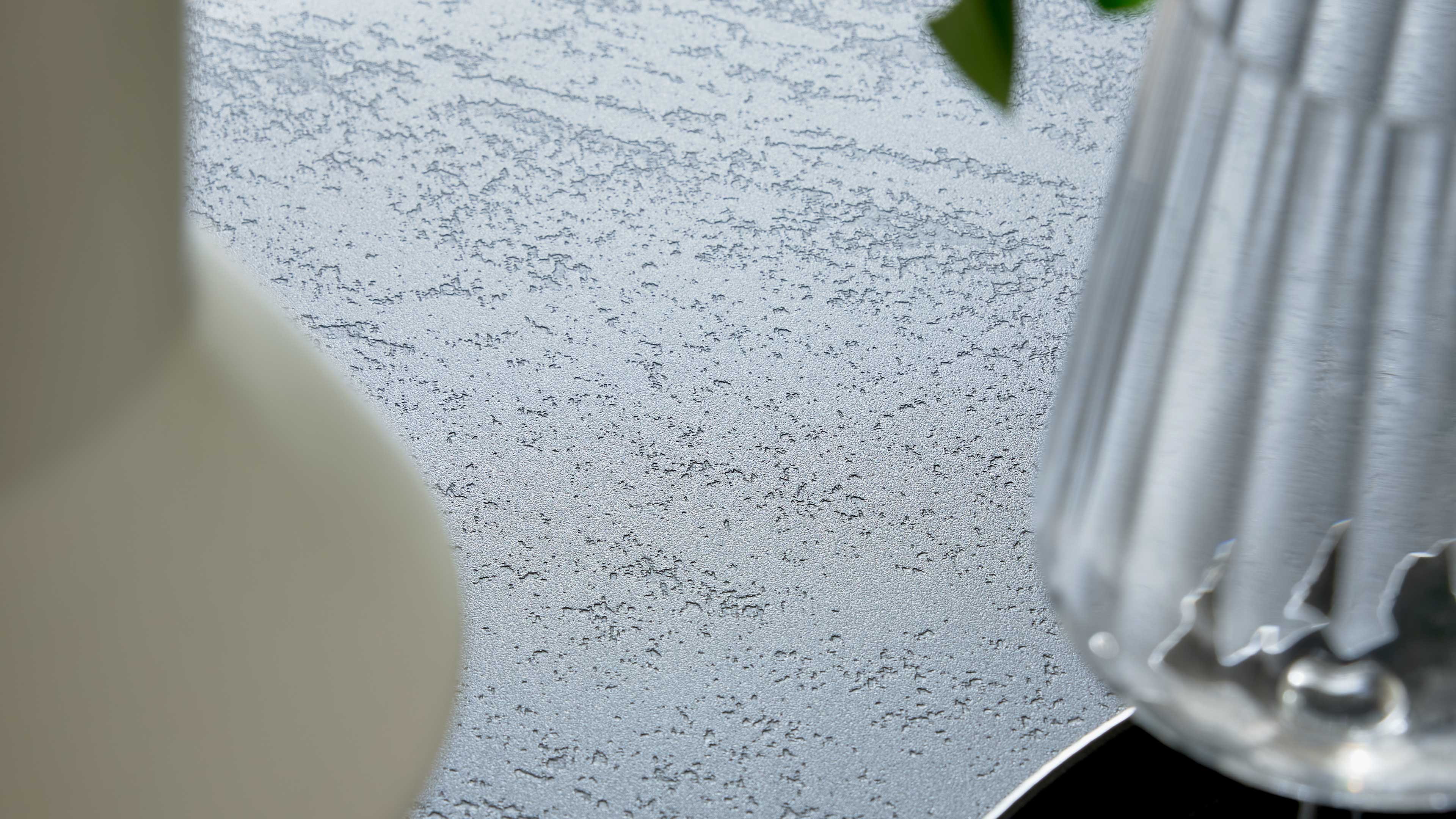 Grau strukturierte Tischplatte mit feiner Körnung, links unscharf eine weiße Vase, rechts ein geripptes Glas.