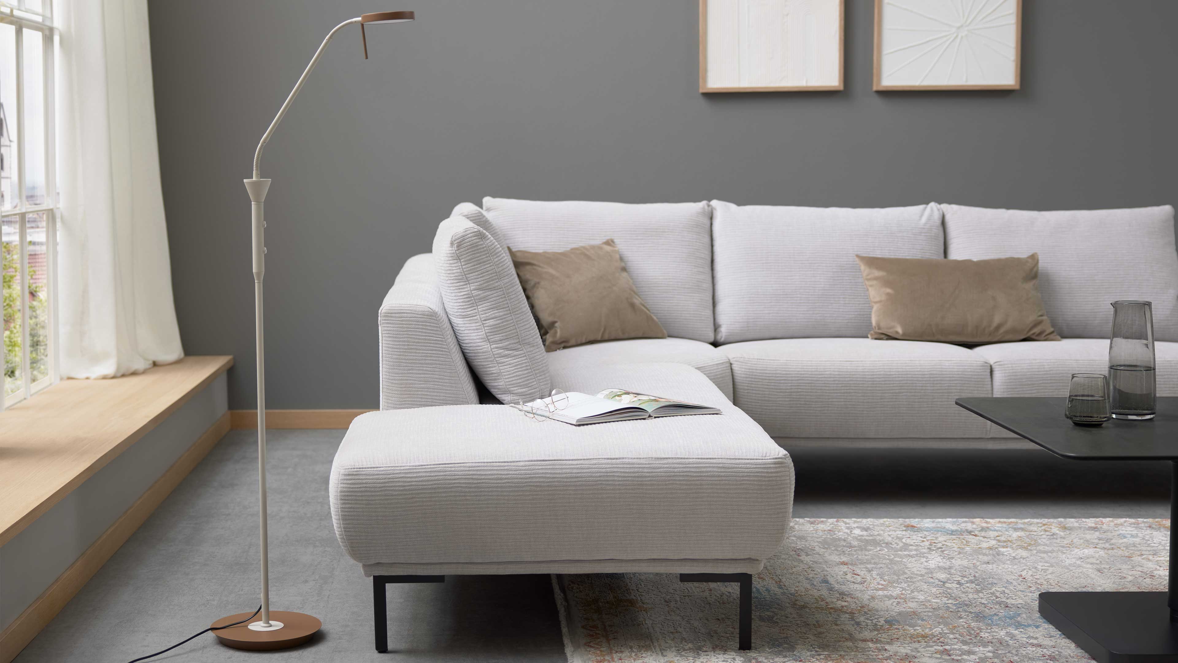 Helles graues Ecksofa mit Chaiselongue, zwei beigen Kissen, offenem Magazin und Brille, Stehlampe links und Couchtisch rechts.