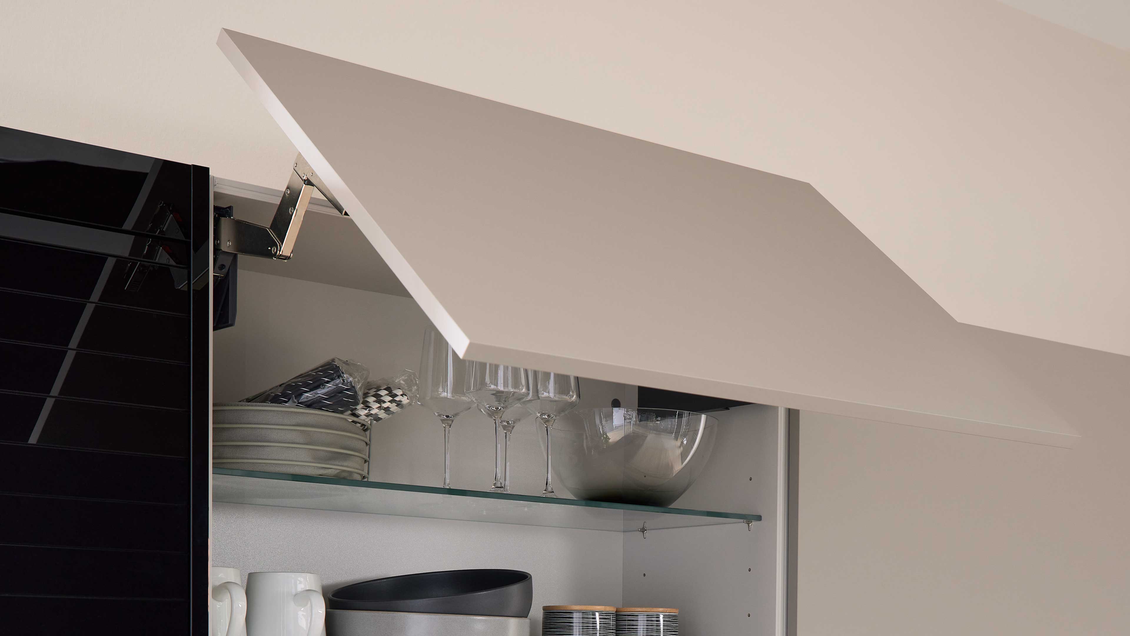 Aufgeklappte helle Oberschrankklappe mit Metallbeschlag, dahinter ein Glasboden mit Weingläsern, Tellern und einer Schüssel.