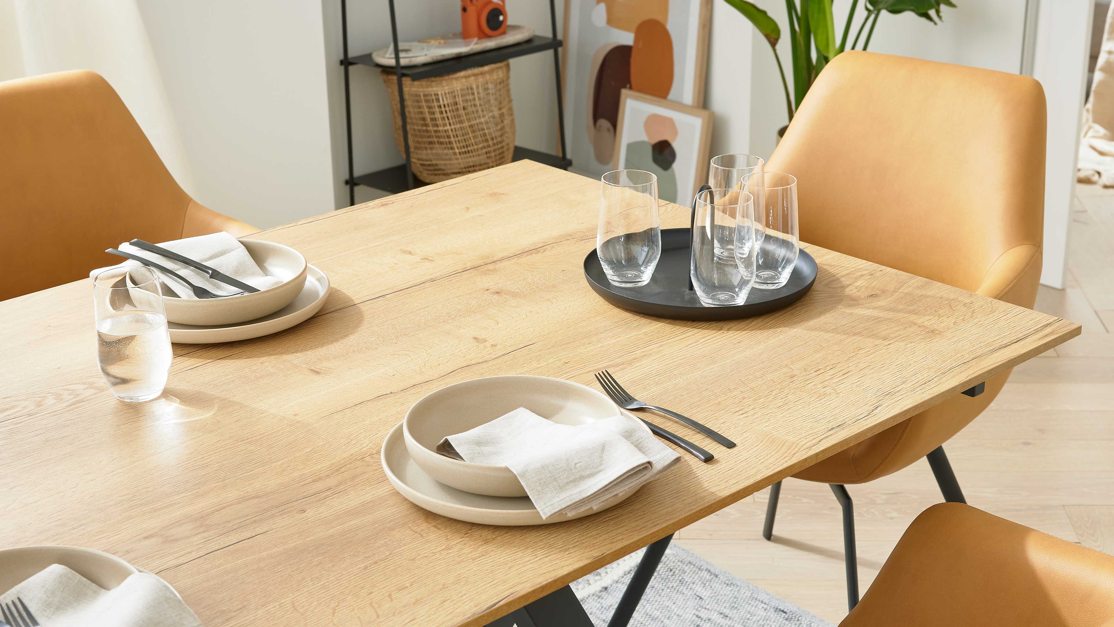 Heller Holztisch mit gedeckten Keramiktellern, Besteck und Stoffservietten, schwarzem Tablett mit vier Wassergläsern und gelben Stühlen.