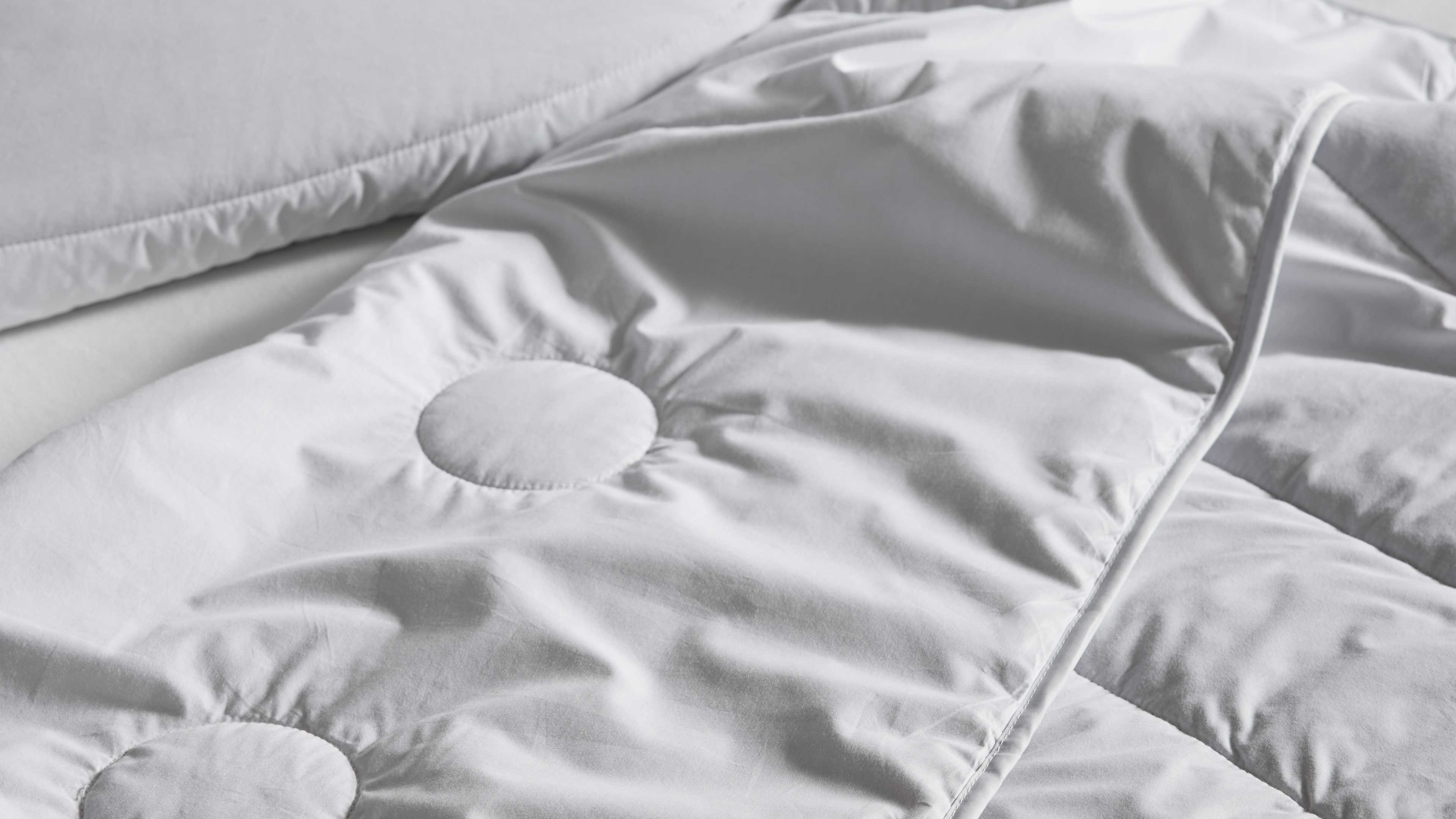Weiß gesteppte Bettdecke mit runden Steppungen, sichtbarer Paspelierung und weichen Falten auf glattem Stoff, links ein Kissenrand.