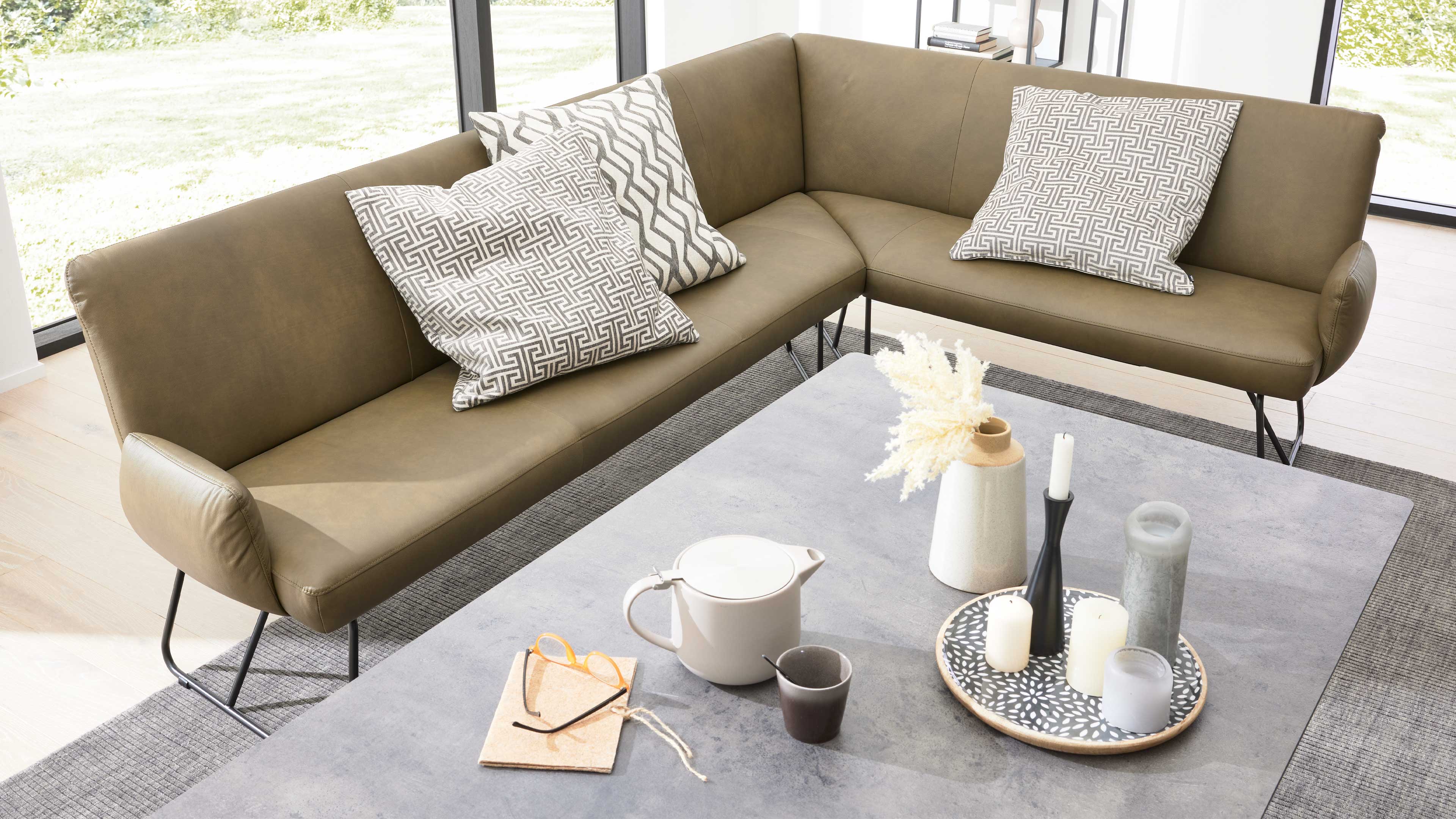 Olivgrünes Ecksofa mit drei gemusterten Kissen, davor Couchtisch mit Teekanne, Tasse, Kerzen, Vase und Notizbuch.