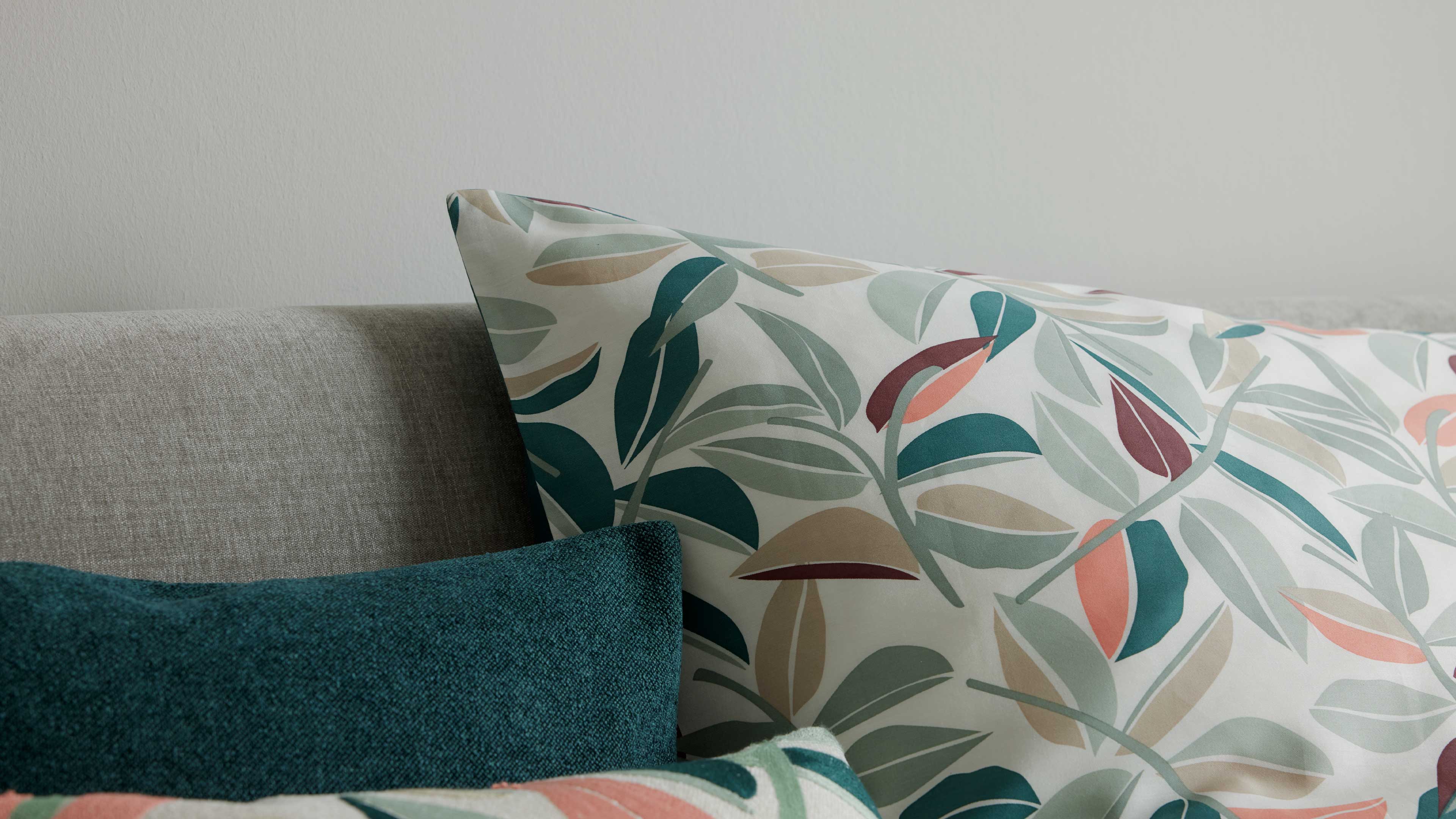 Großes Kissen mit pastellfarbenem Blattmuster liegt auf einer hellgrauen Sofalehne, davor ein grüntürkis-texturiertes Kissen.