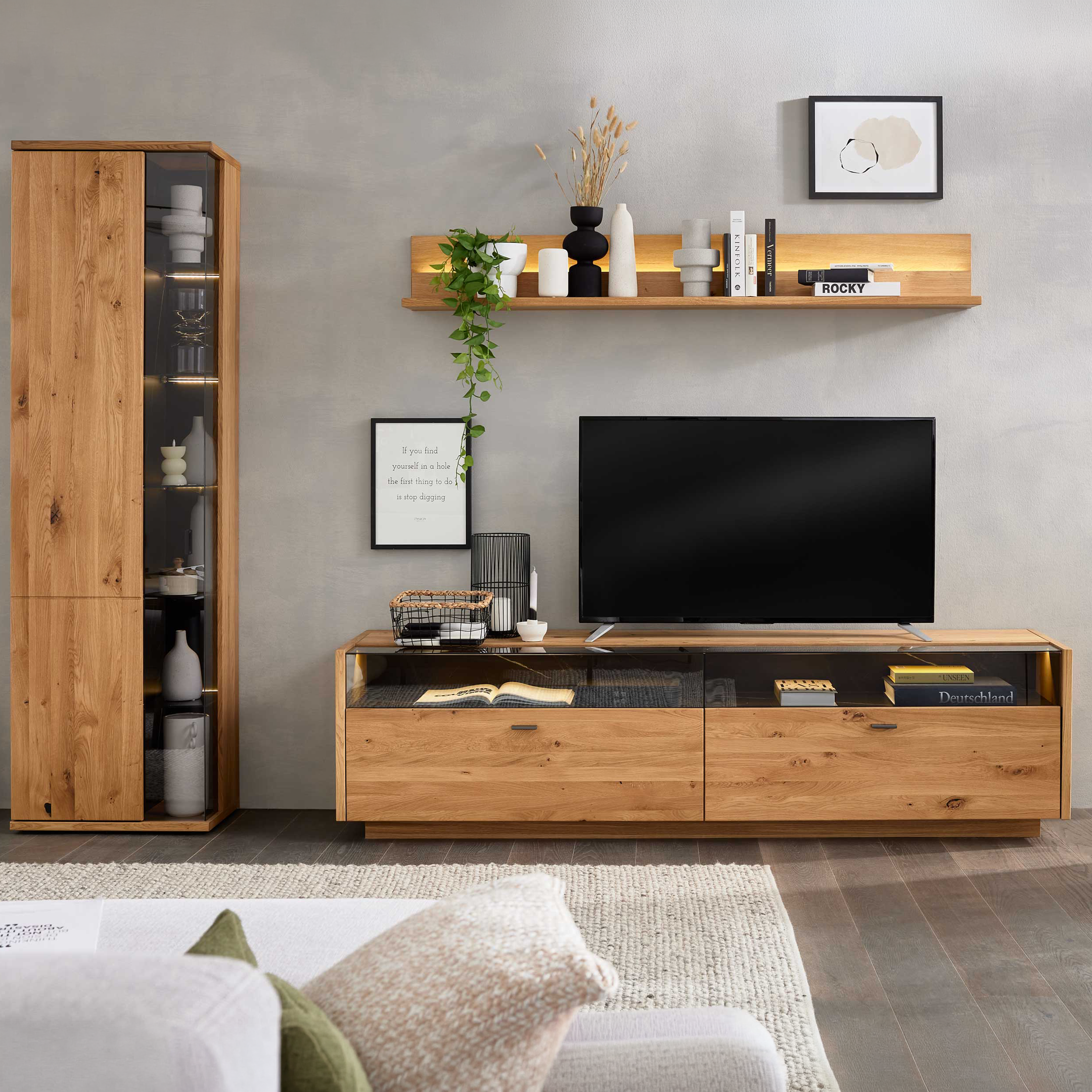 Helles Holz-Medienboard mit Flachbildschirm vor einer dunkelgrünen Wand, daneben hängender Glasschrank und Pflanze.