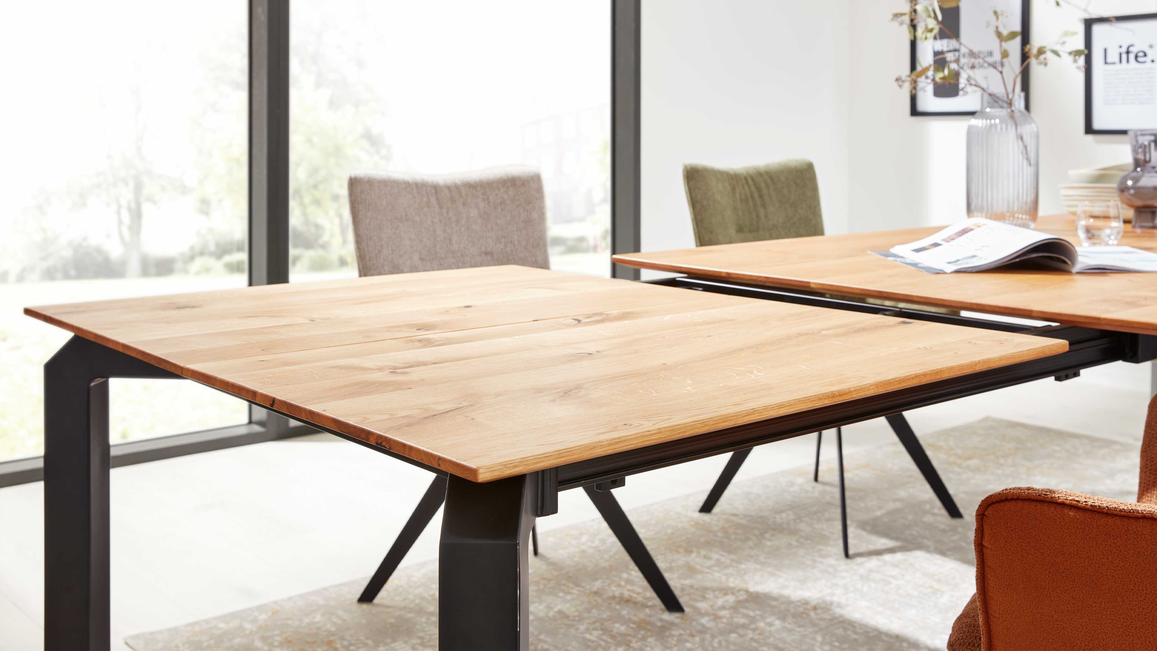 Großer Esstisch mit heller zweigeteilter Holzplatte auf schwarzen Metallbeinen, sichtbare Auszugsführungen und mehrere gepolsterte Stühle.