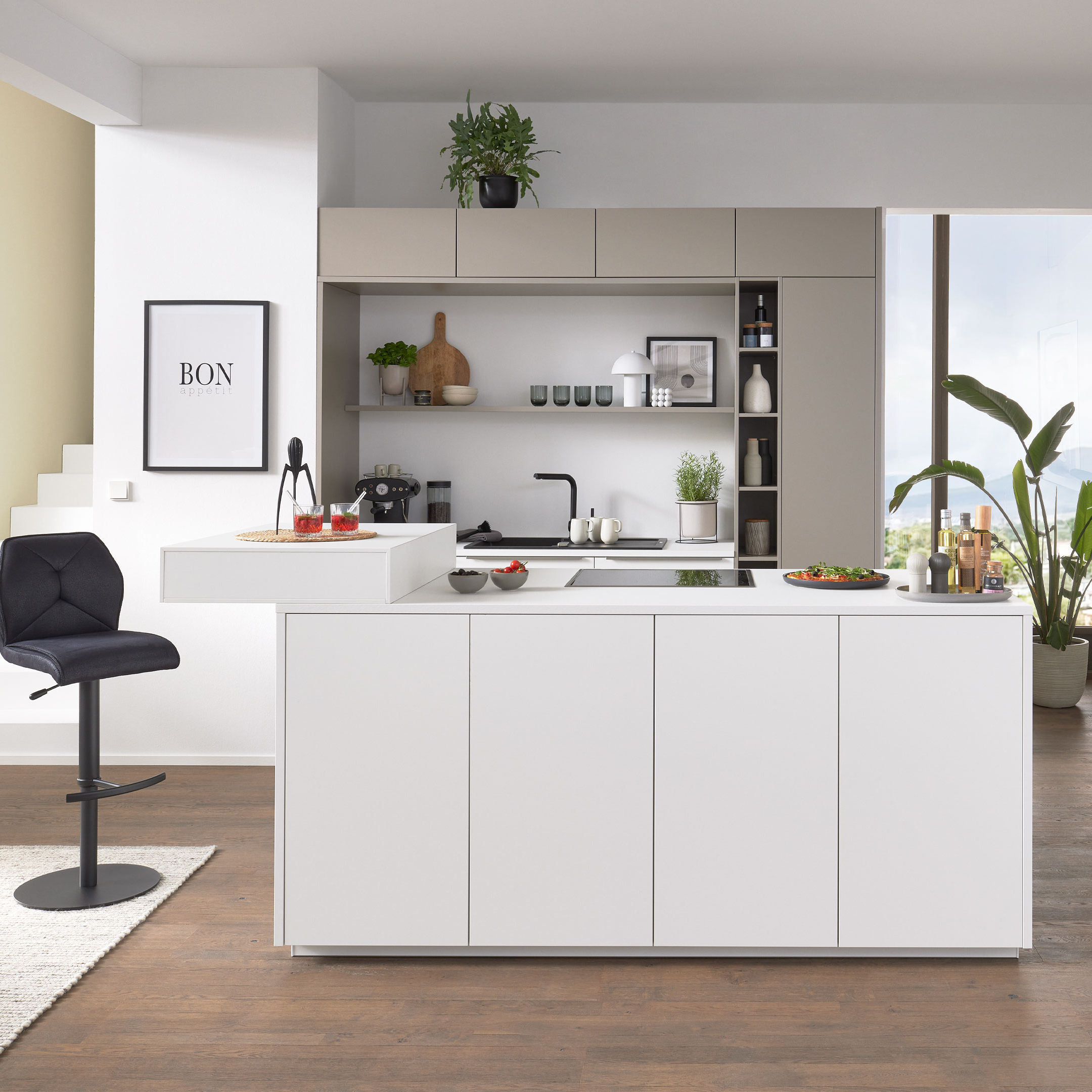 Zentrale weiße Kücheninsel mit Kochfeld und erhöhtem Tresen, dahinter beige Hochschränke, offenes Regal und schwarzer Barhocker.