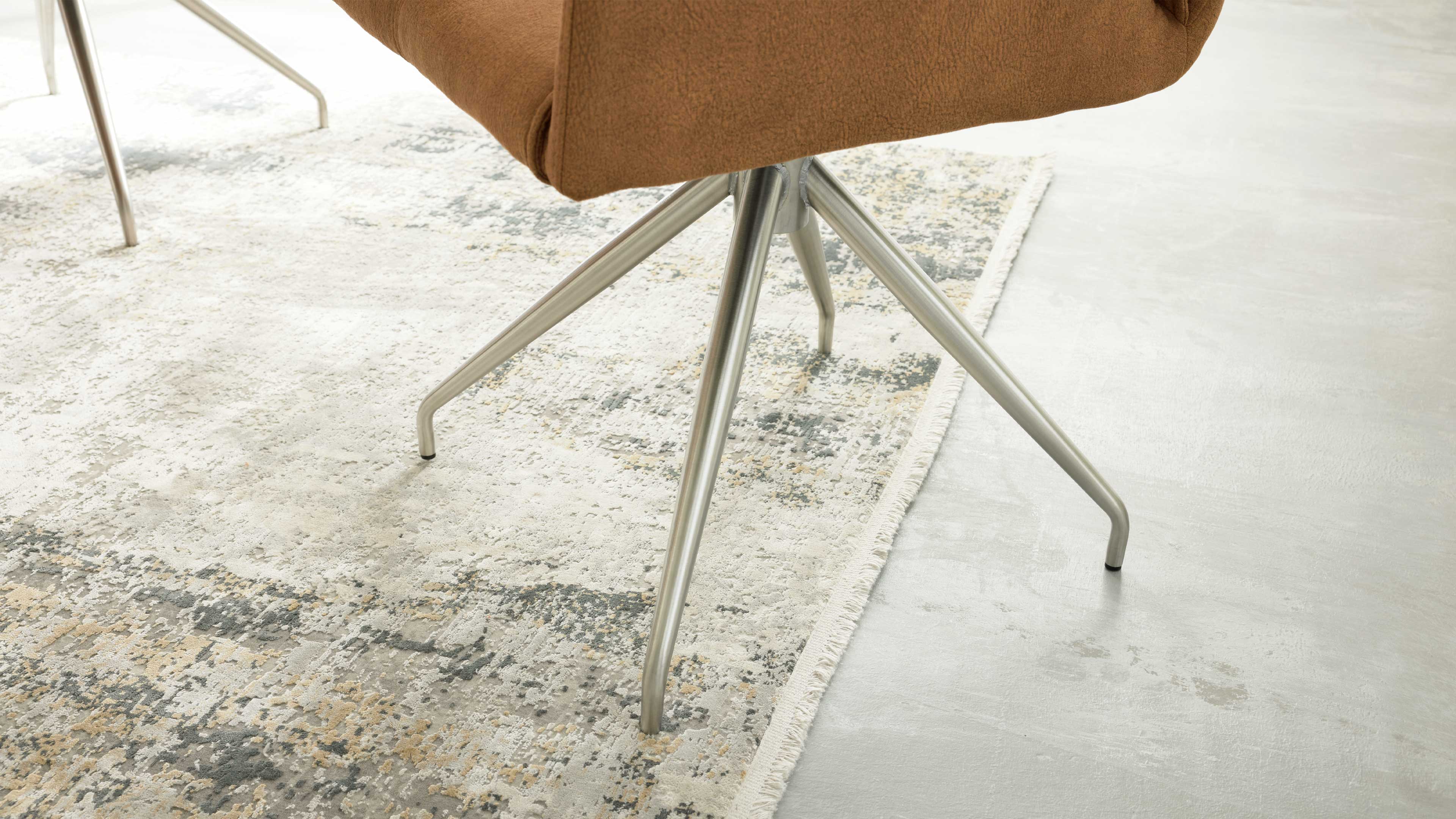 Brauner Polsterstuhl auf vierarmigem, gebogenem Metallfuß, teilweise auf einem hell gemusterten Teppich mit Fransen.