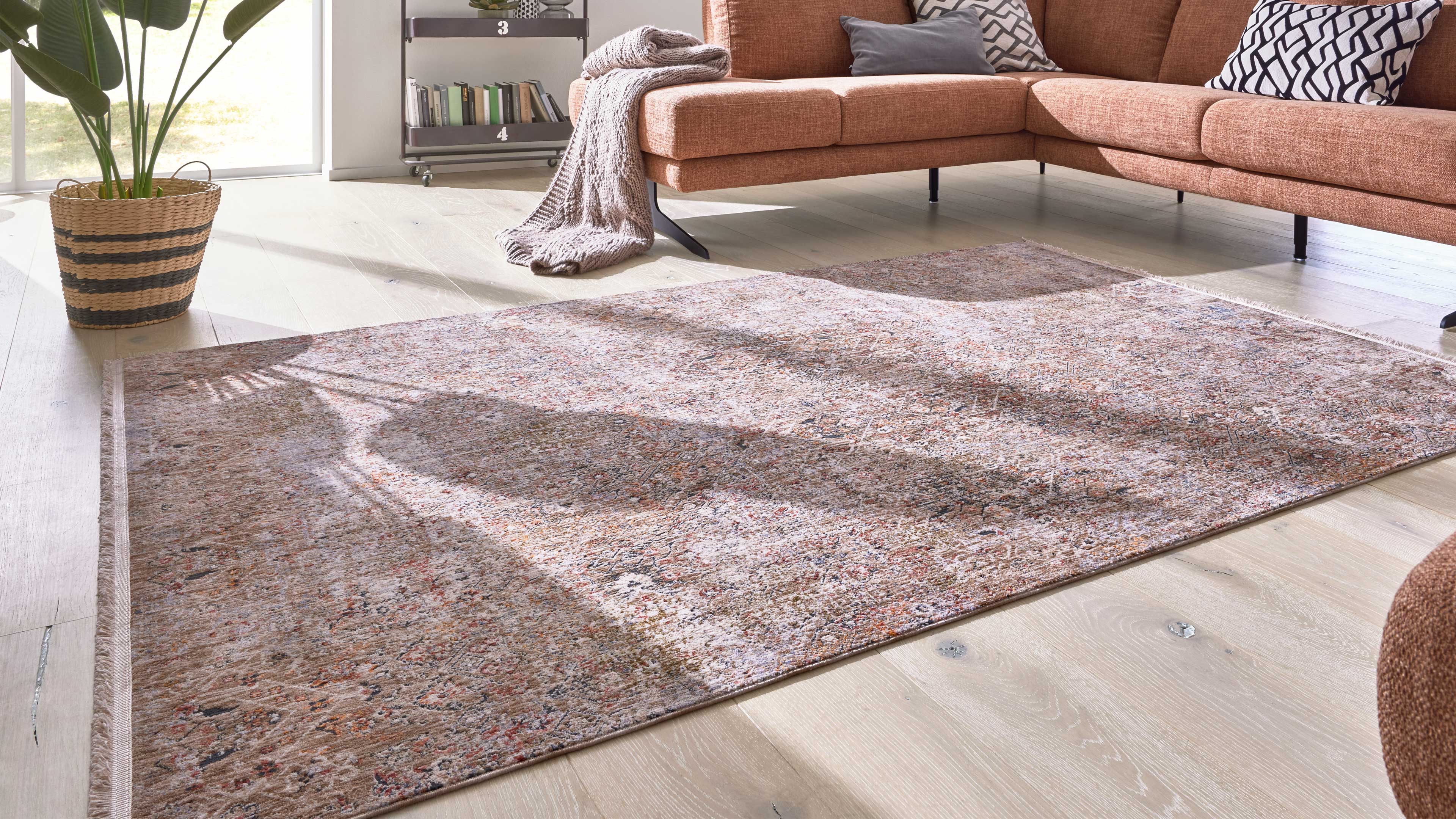 Großer Teppich mit dezenter Musterung auf hellem Holzboden, daneben rostfarbenes Ecksofa, Korb mit Pflanze und Decke.