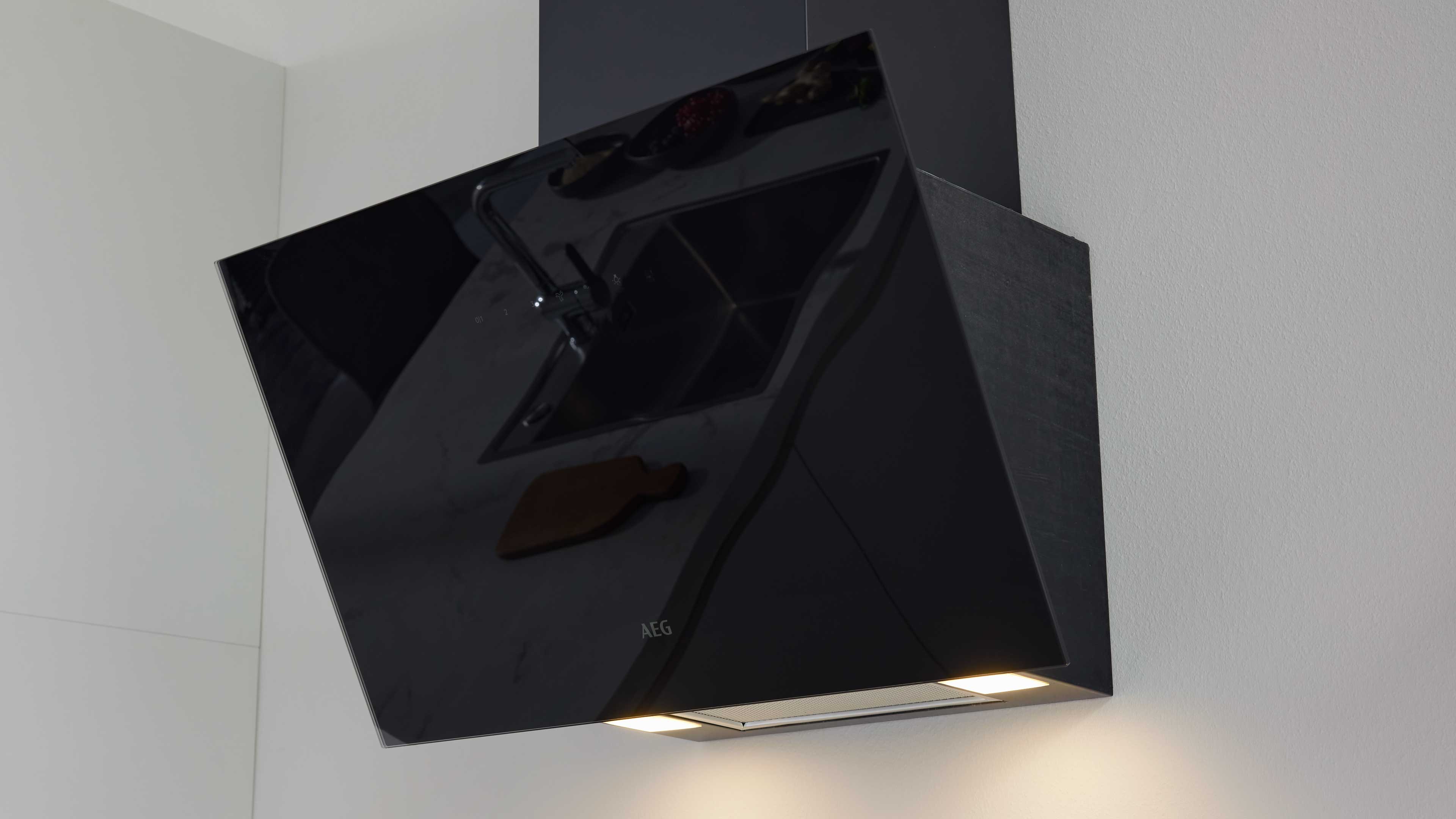Schwarze, schräg an der Wand montierte Dunstabzugshaube mit spiegelnder Front, sichtbarer Spülenreflexion und eingeschaltetem Licht.