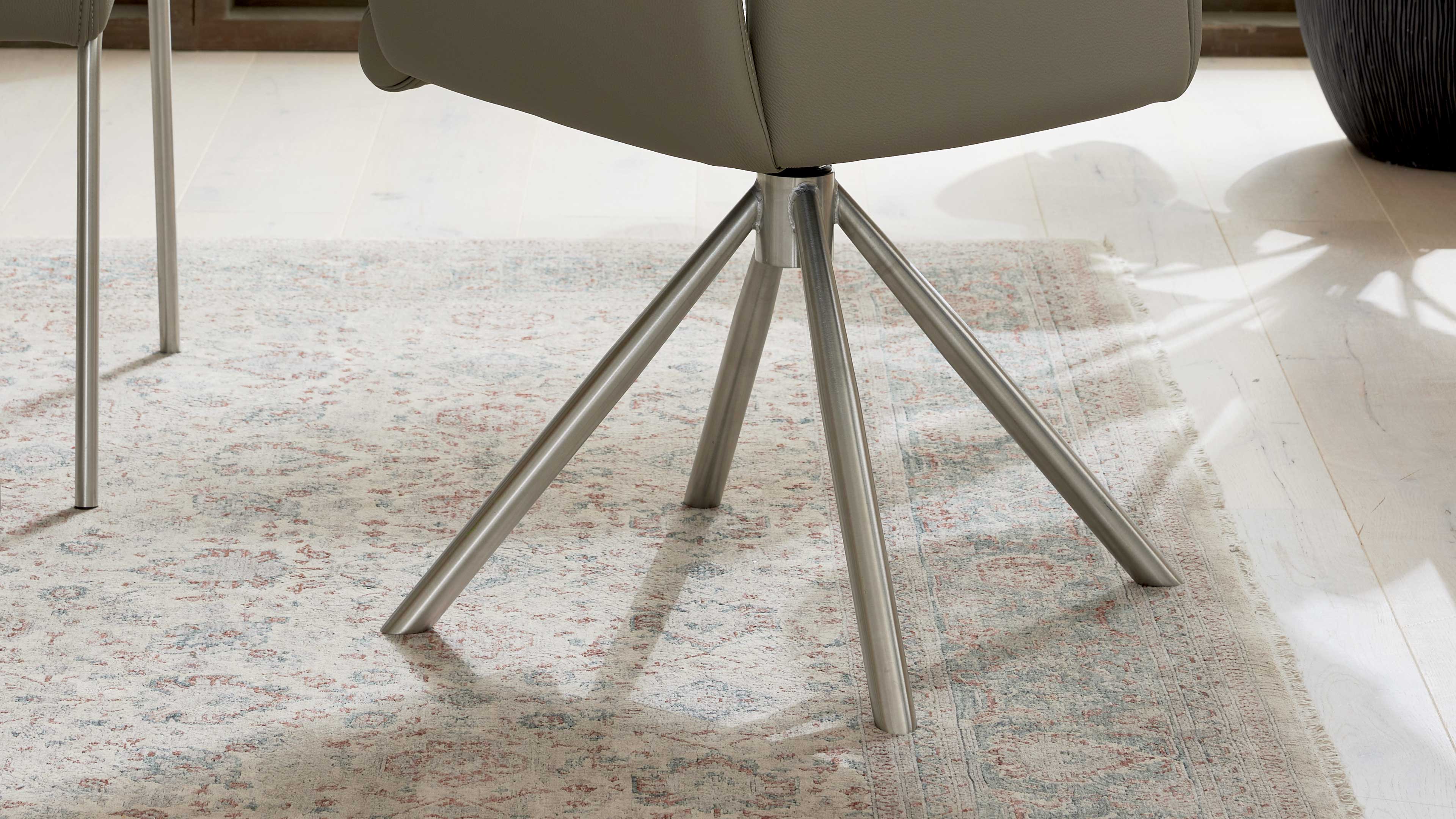 Sternförmiger Metallfuß eines Stuhls mit schräg abgestellten Rohren auf hell gemustertem Teppich und hellem Holzboden.