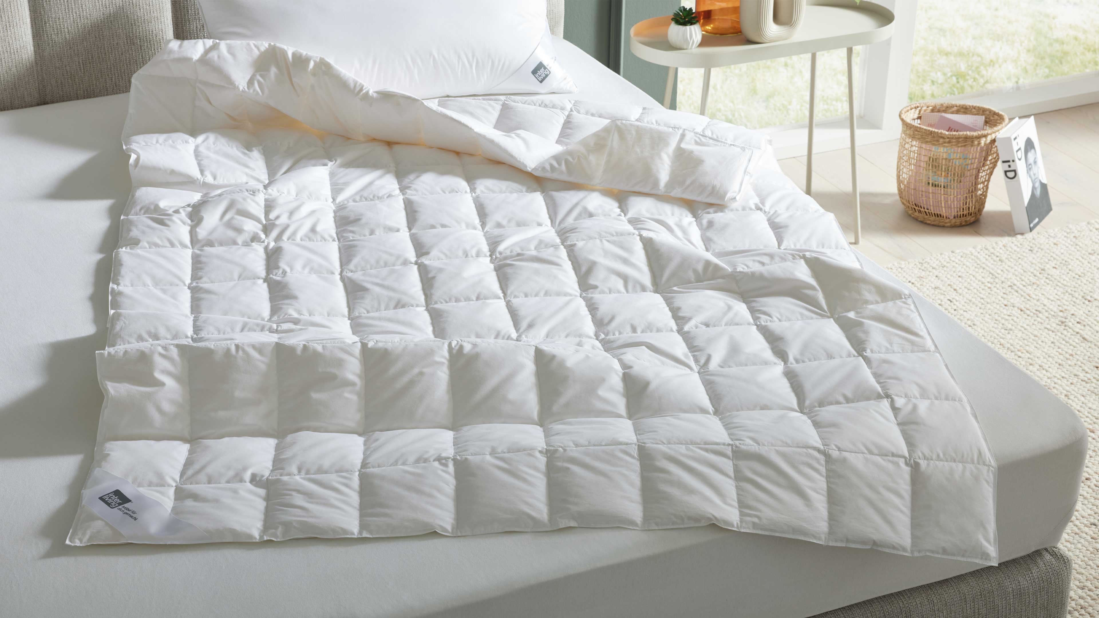 Weiße gesteppte Bettdecke liegt teilweise aufgefaltet auf Matratze, mit weißem Kopfkissen, Beistelltisch und Korb daneben.