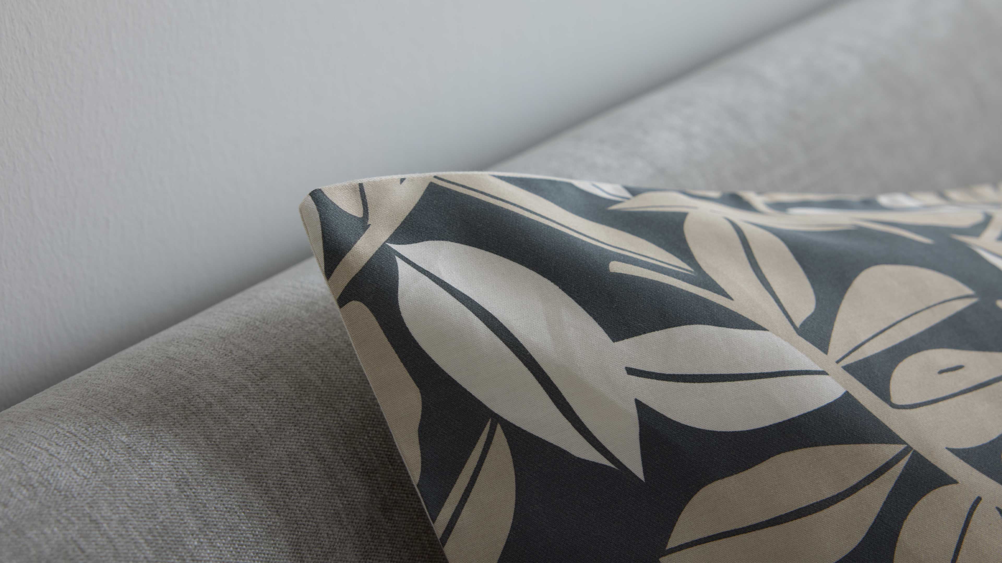 Sofakissen mit großem beige-blättrigem Muster auf dunklem Stoff, liegend auf grauem Sofa vor heller Wand.