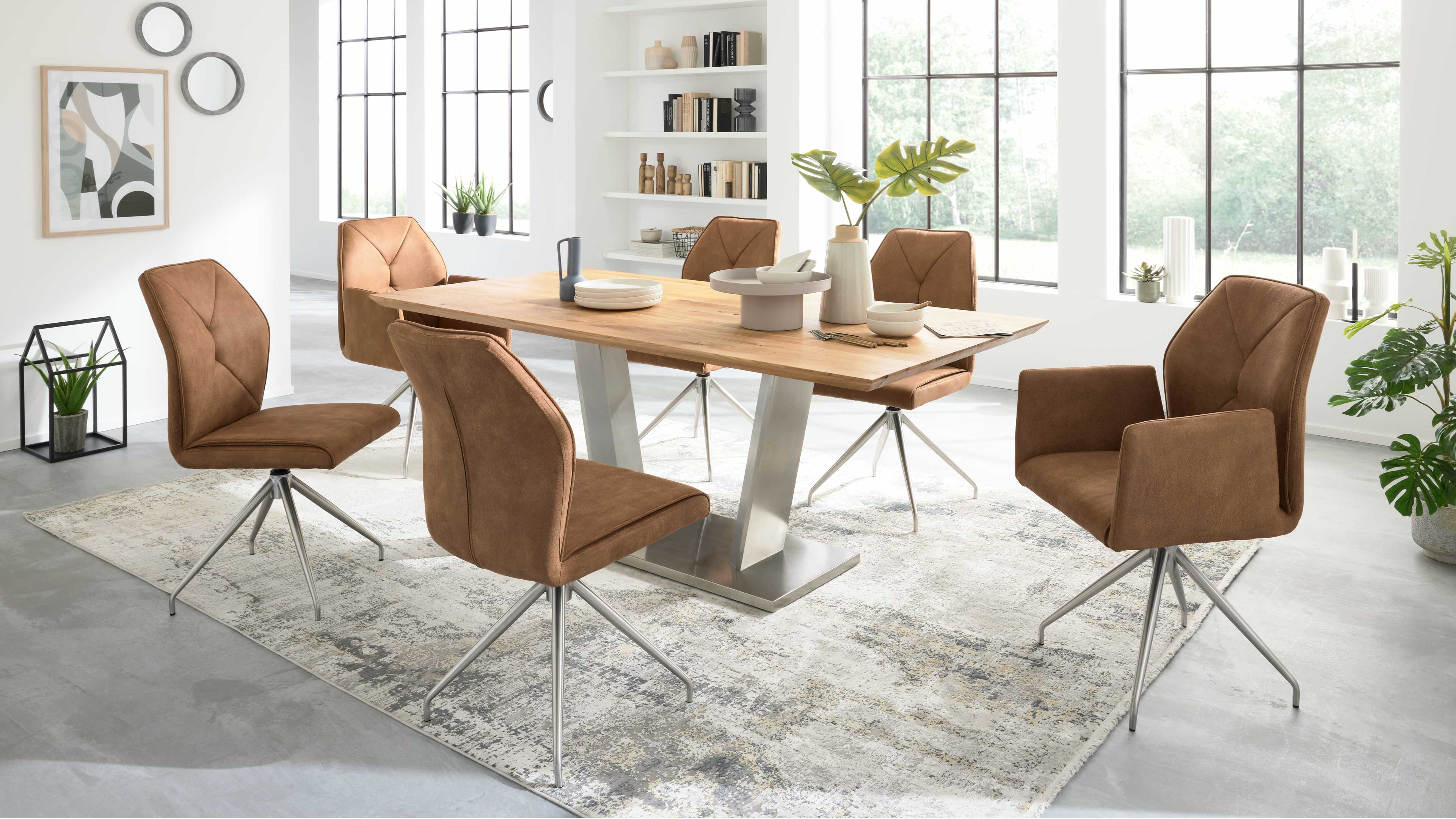Esstisch aus hellem Holz auf silbernem V-Fuß, umgeben von sechs braunen, gepolsterten Drehstühlen mit Metallgestell.
