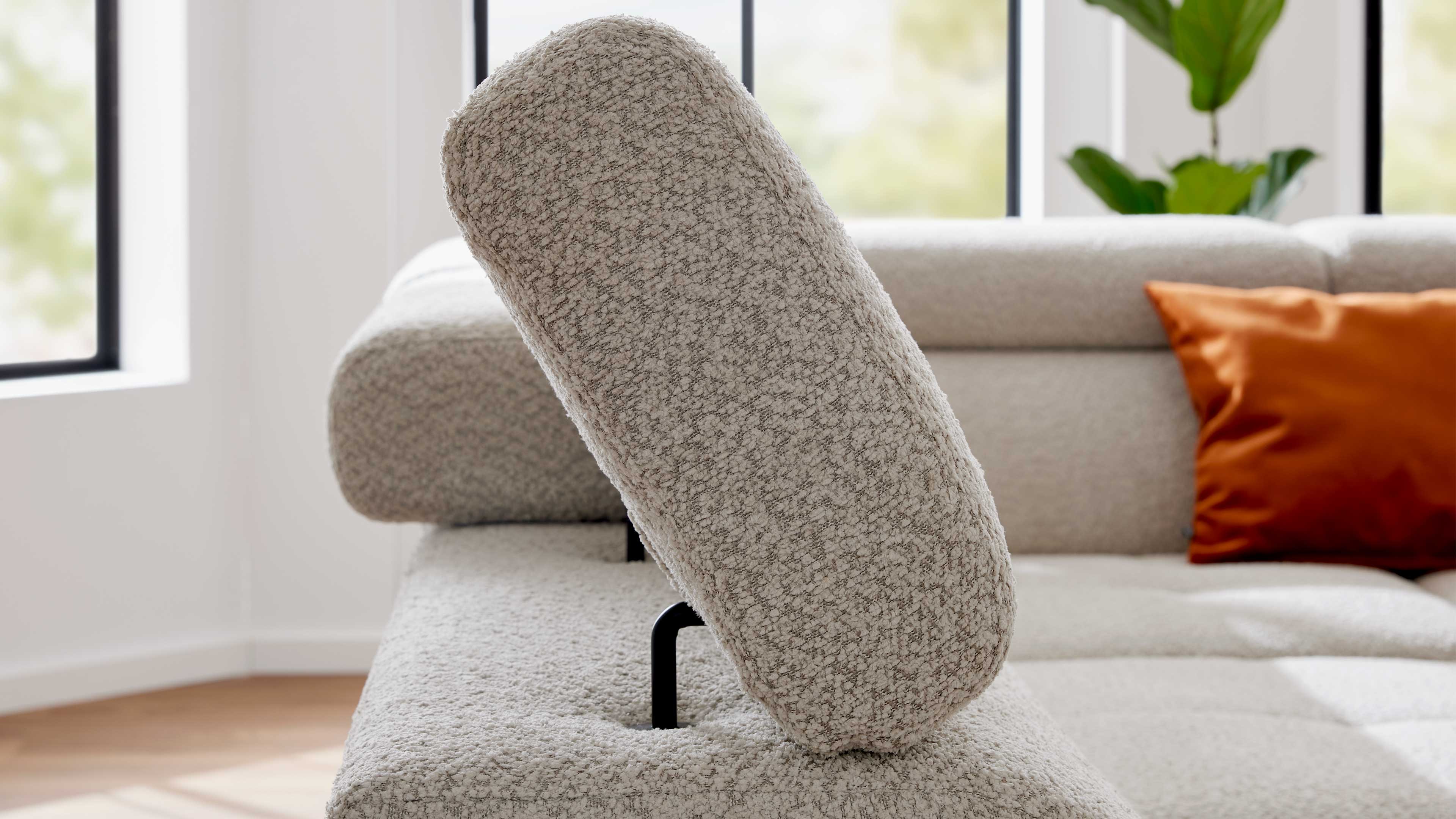 Verstellbare, beige texturierte Polsterlehne auf Sofa mit sichtbarem schwarzen Stützbügel, im Hintergrund Sofa und orangefarbenes Kissen.