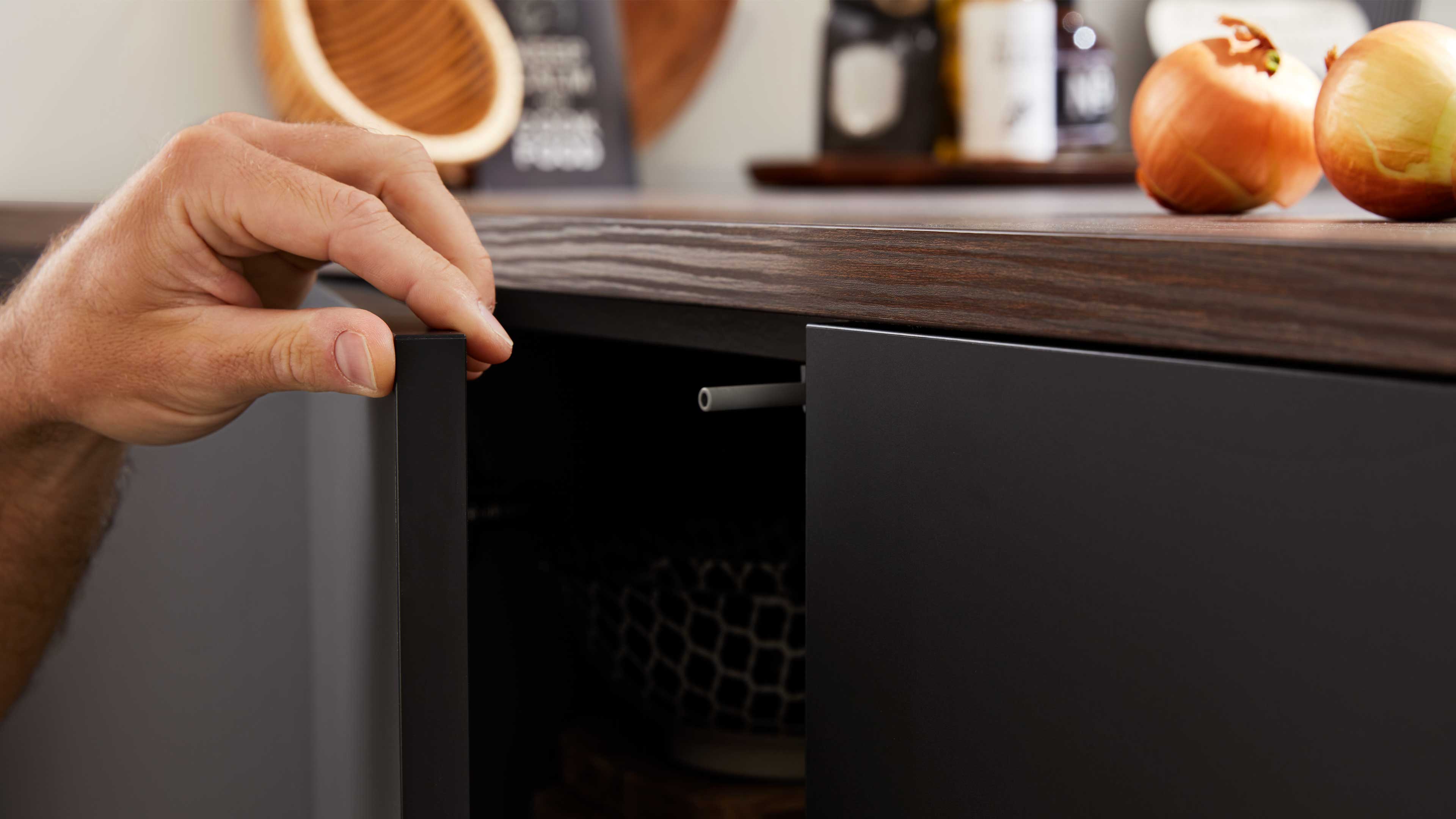 Hand öffnet matte dunkelgraue Schranktür unter einer dunkel gemaserten Holzarbeitsplatte; auf der Platte zwei Zwiebeln und unscharfe Deko.