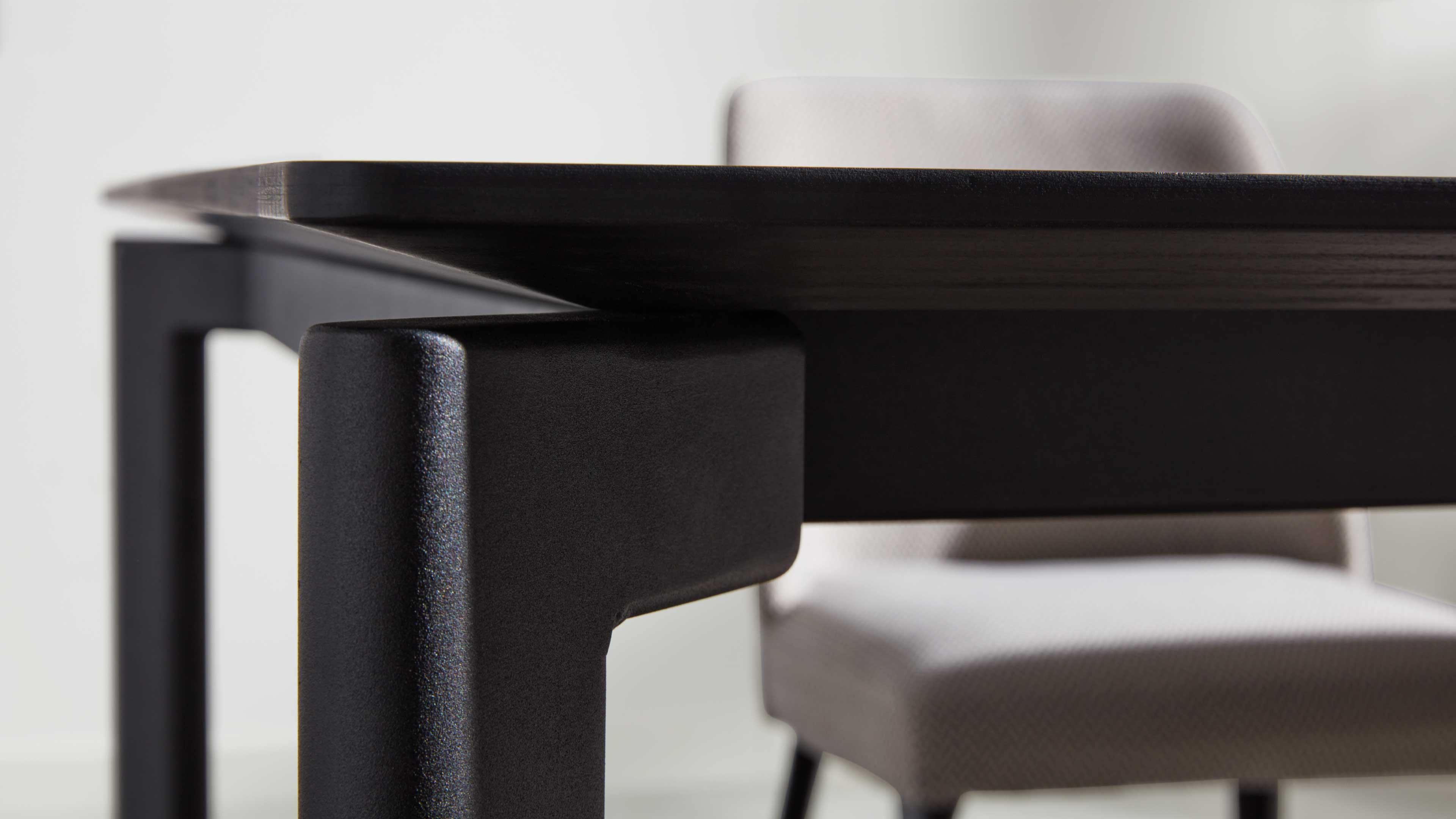 Schwarzes, mattes Tischgestell mit überstehender Tischplatte und abgerundeter Ecke, im Hintergrund ein heller gepolsterter Stuhl.