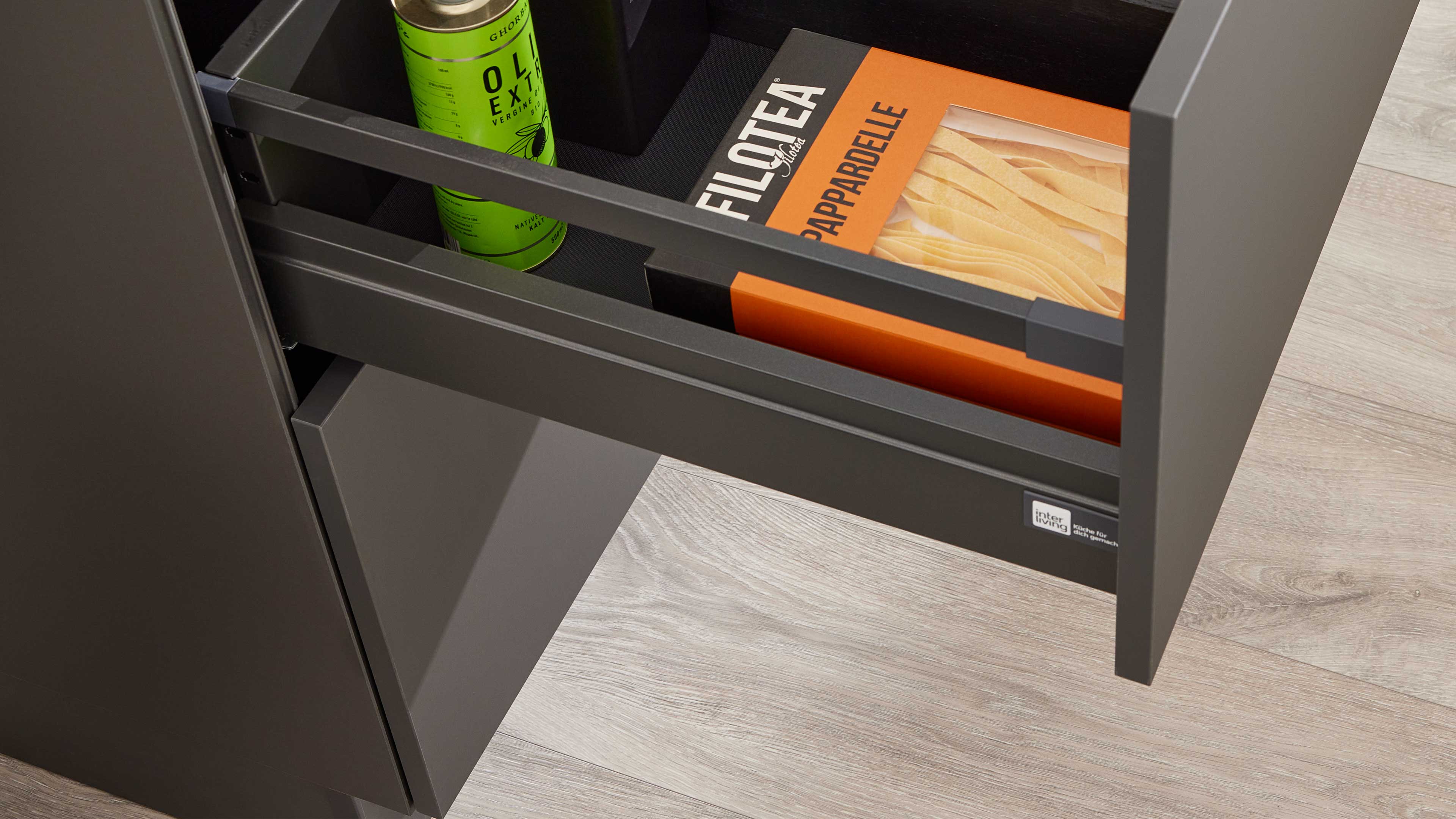 Geöffnetes graues Küchenschubfach mit Metallleisten, darin ein orangefarbener Pastakarton und eine grüne Ölkanne auf hellem Holzfußboden.