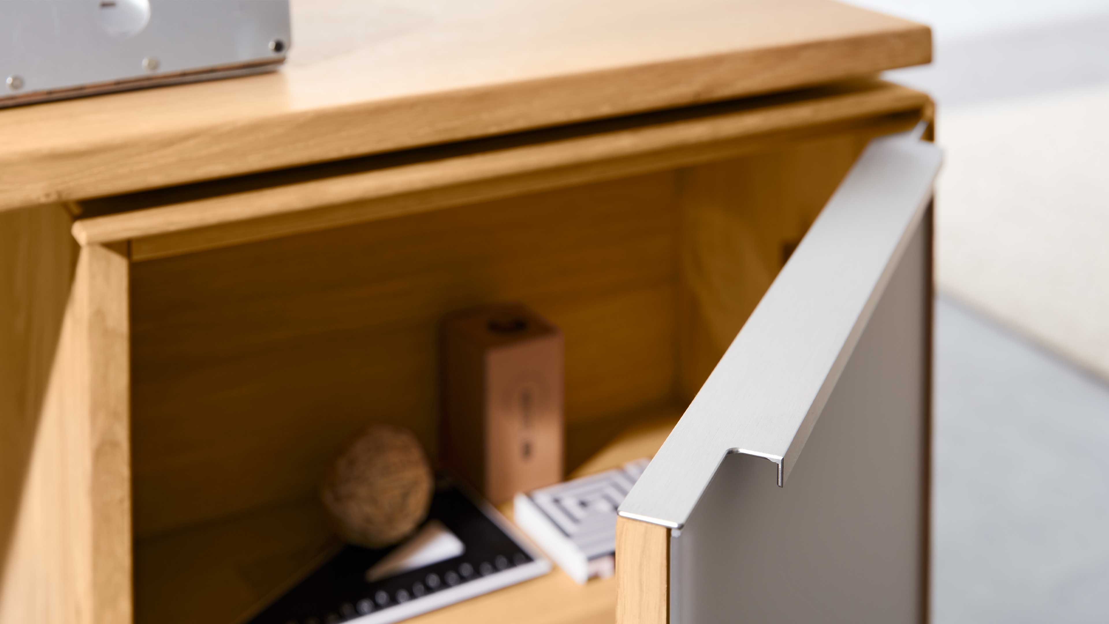 Holzschrank mit geöffneter Tür und silberner Griffleiste, im Inneren eine Deko-Kugel, eine braune Schachtel und zwei Notizbücher.