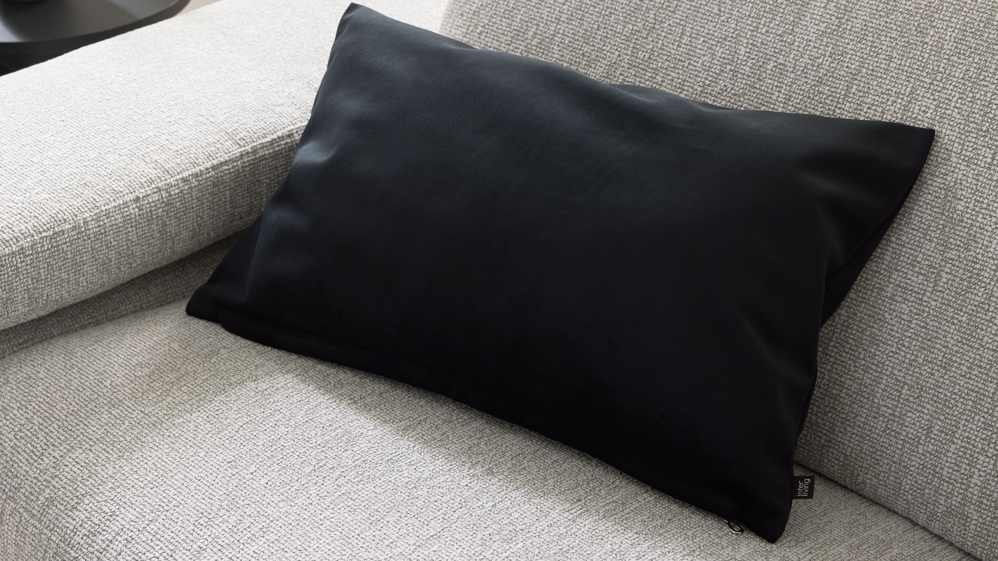 Schwarzes rechteckiges Kissen mit sichtbarem Reißverschluss und Stoffetikett liegt schräg auf hellgrauem, strukturiertem Sofa.