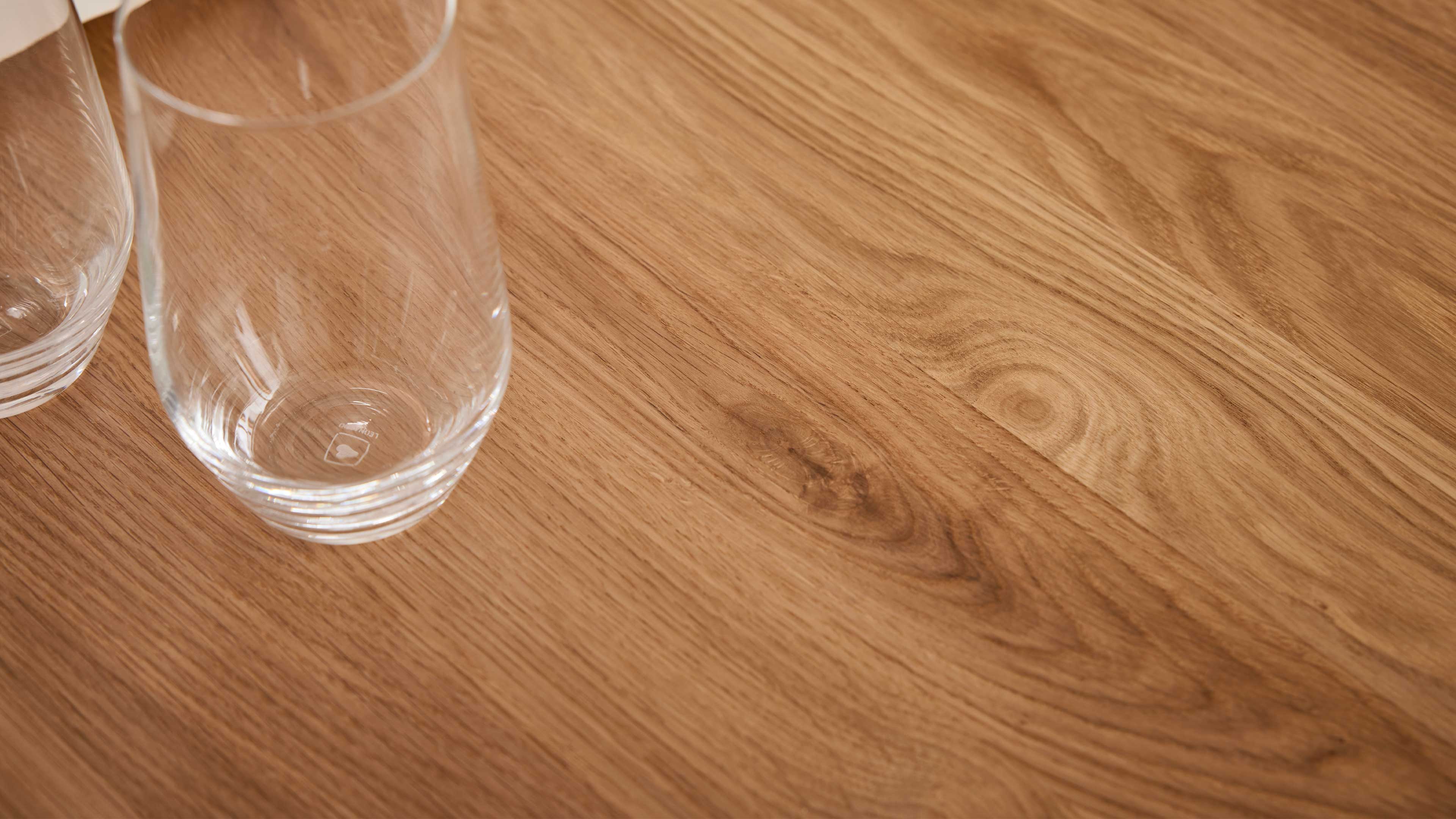 Helle Holz-Tischfläche mit sichtbarer Maserung und zwei teilweise sichtbaren, klaren Gläsern am linken Bildrand.