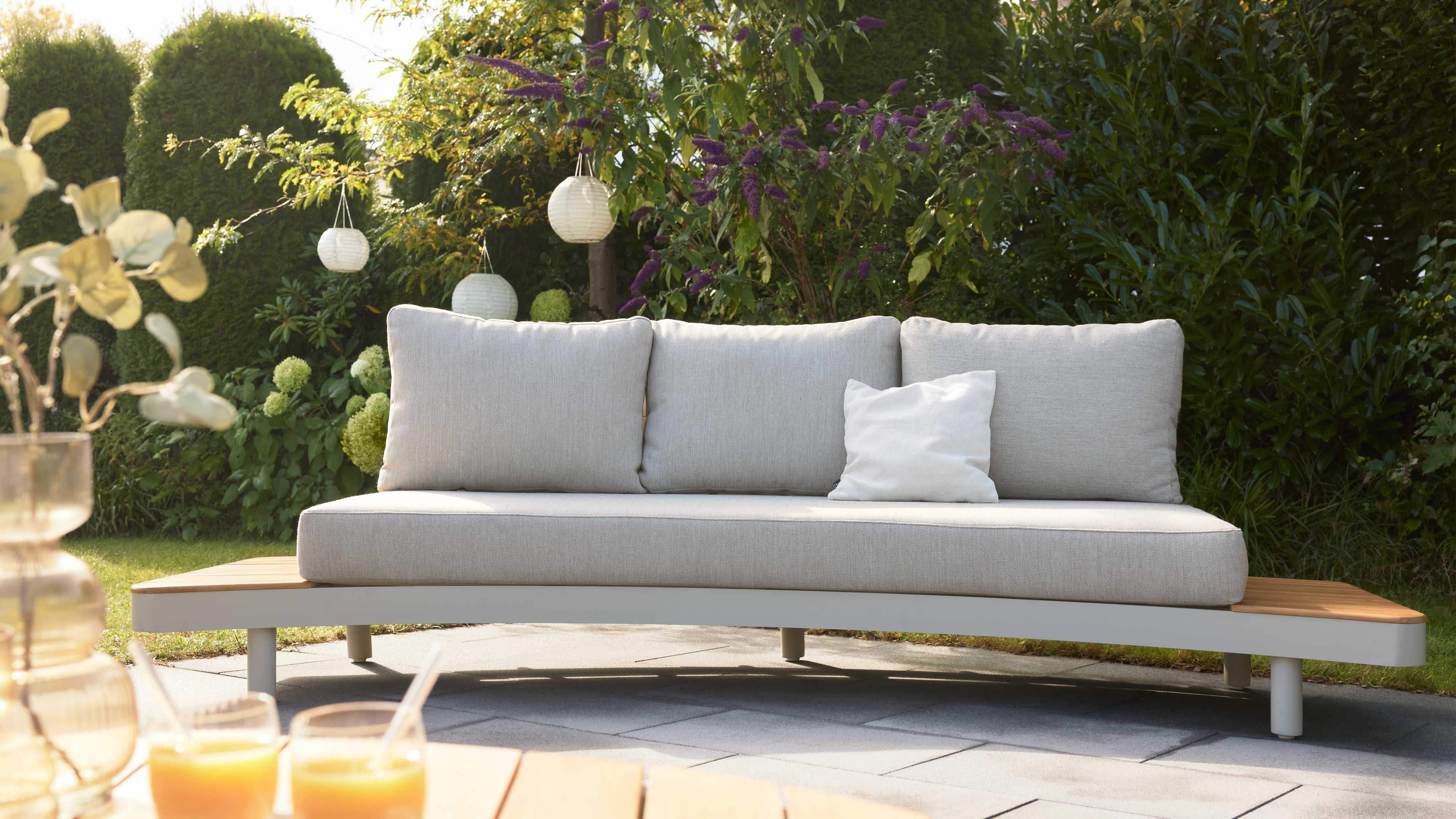 Hellgraues Sofa mit drei großen Rückenkissen und einem weißen Kissen, Holzseitenteilen, auf gepflasterter Terrasse vor grüner Hecke.