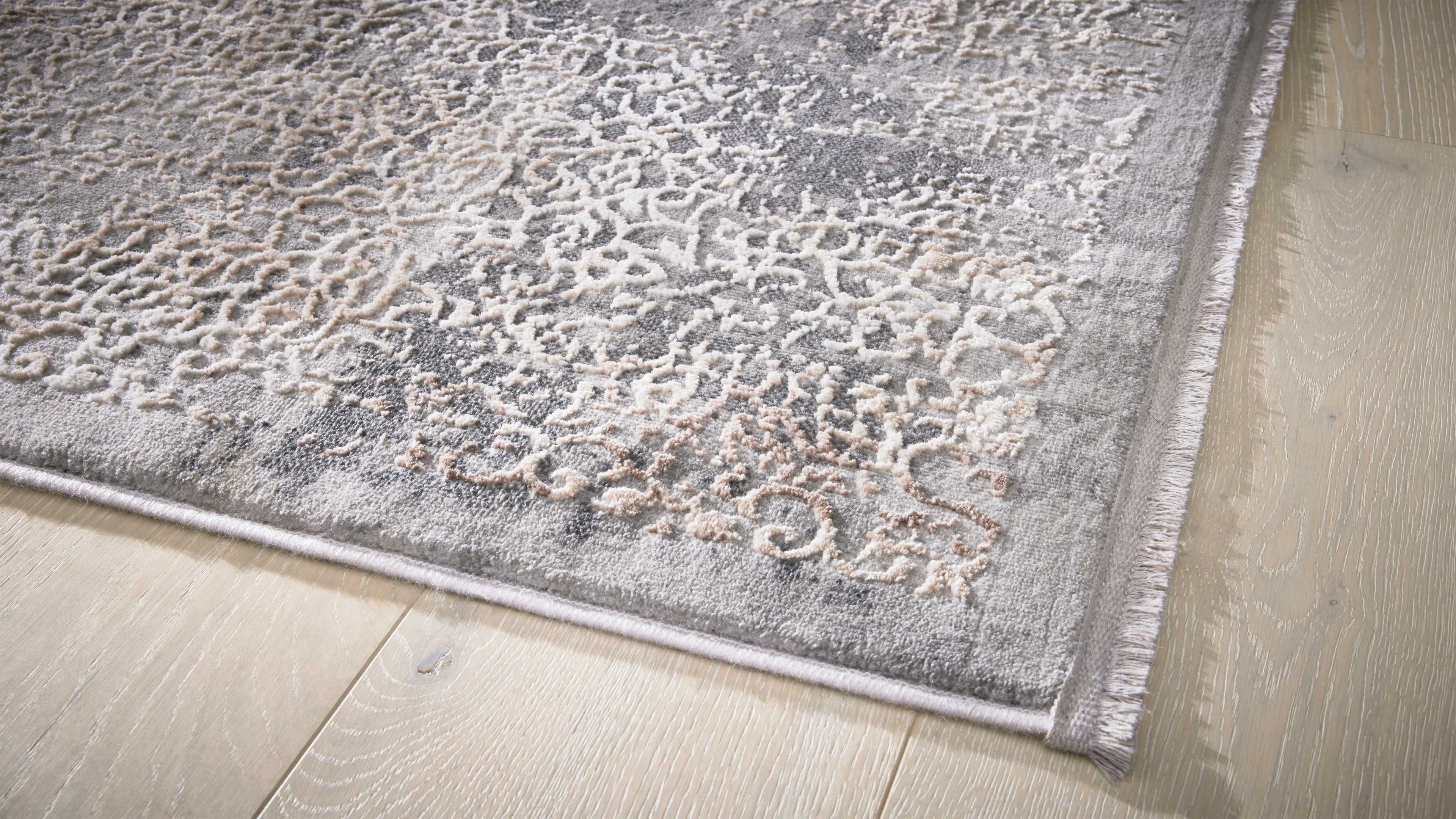 Grauer Teppich mit hellbeigem Reliefornament, eingefasster Kante und Fransen rechts auf hellem Dielenboden.