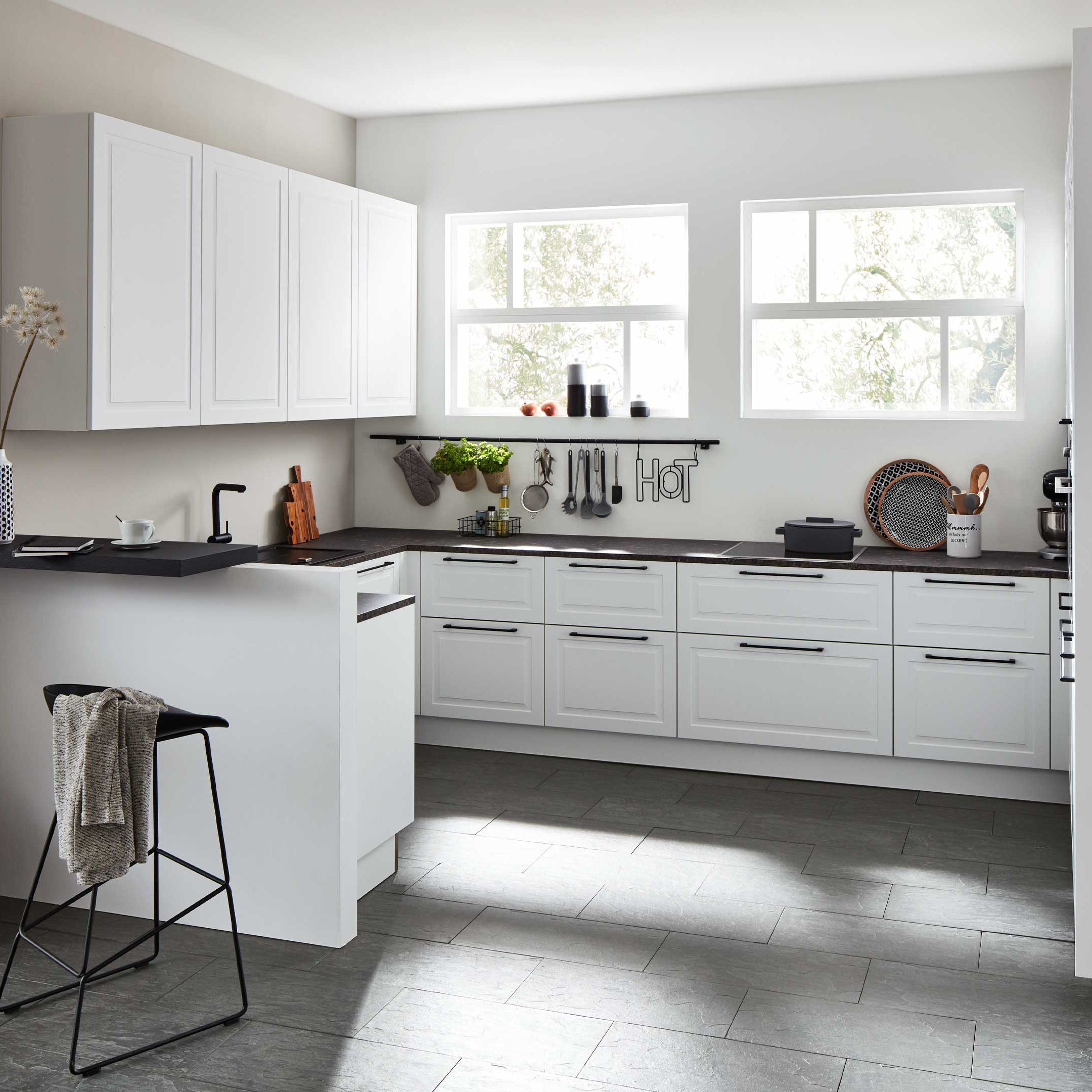 Weiße Küchenunterschränke und Hängeschränke mit dunkler Arbeitsplatte, zwei Fenster, Hakenleiste mit Utensilien und ein Barhocker.