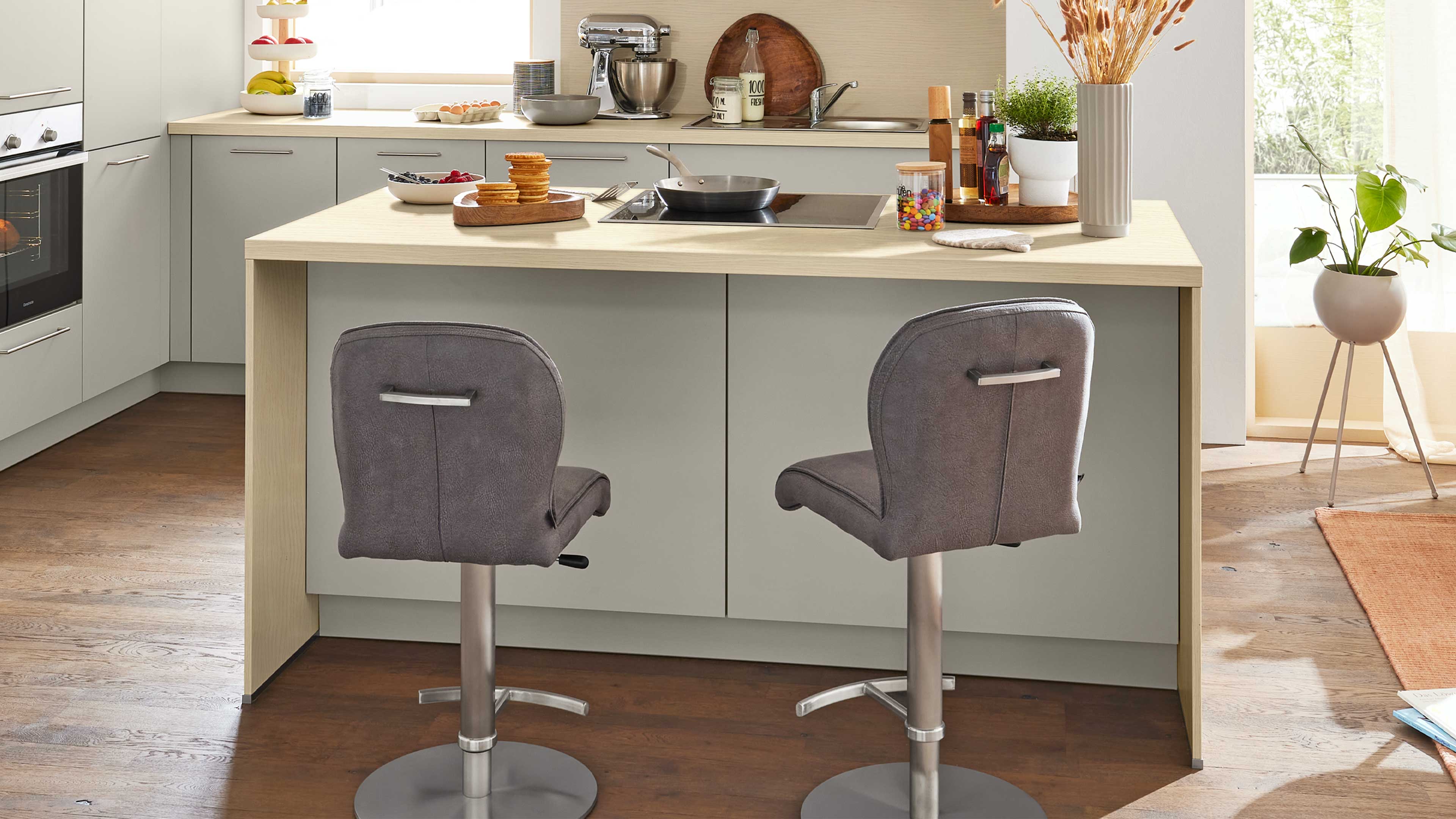 Zwei graue, höhenverstellbare Barhocker stehen vor einer hellen Kücheninsel mit Holzarbeitsfläche, Kochfeld und Deko.